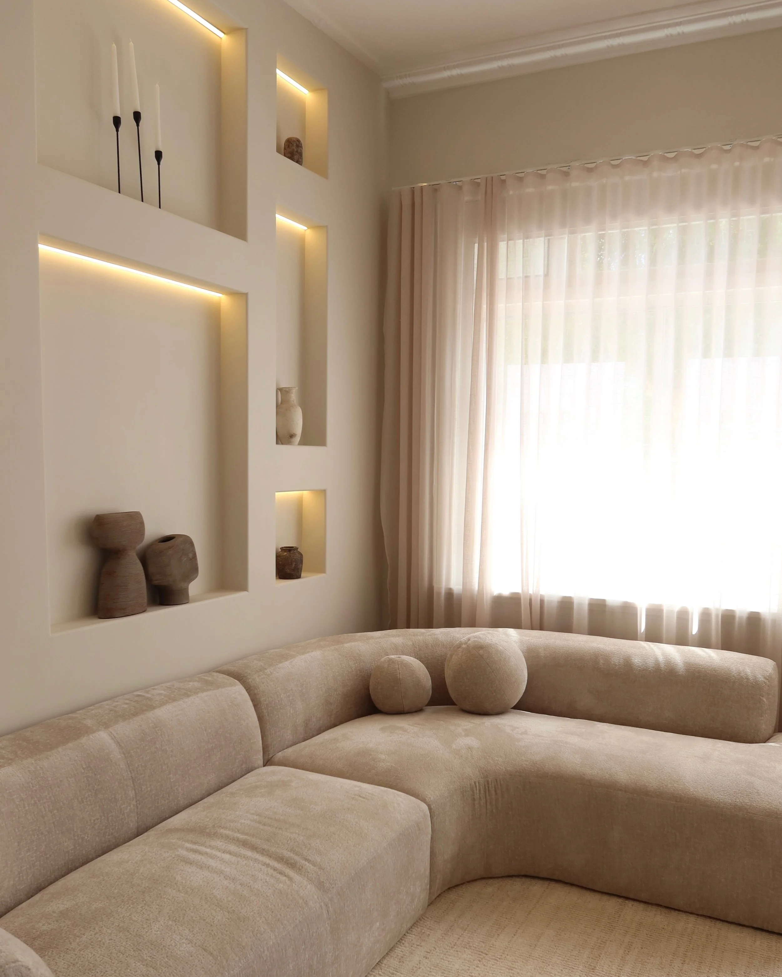 Living room corner with beige sectional sofa, two round pillows, a wall with built-in display niches with decorative items, and a large window with sheer curtains.
