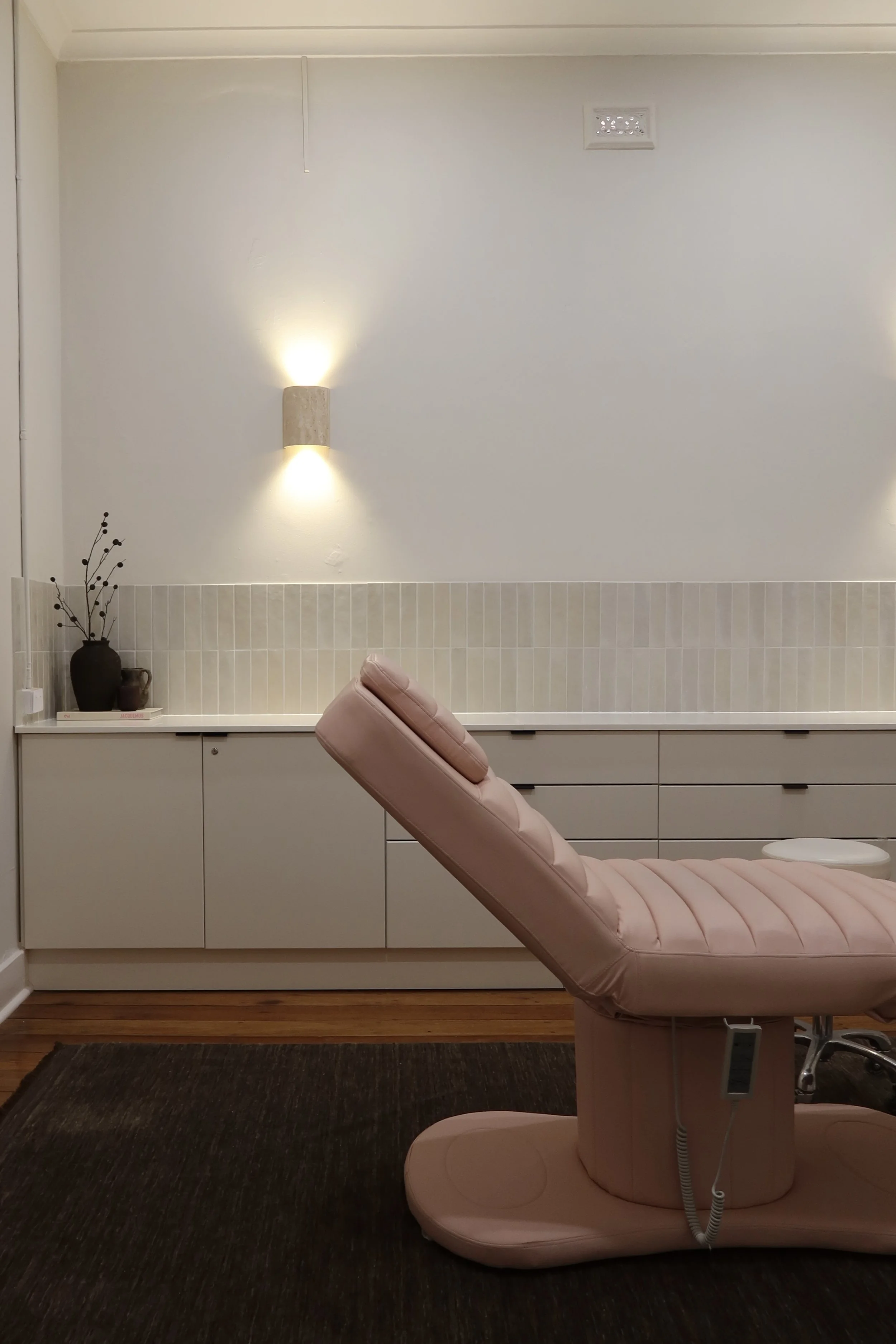 A pink medical or dental examination chair in a clean, modern room with white cabinets, beige tile backsplash, a black vase with dried branches, and wall-mounted lights.