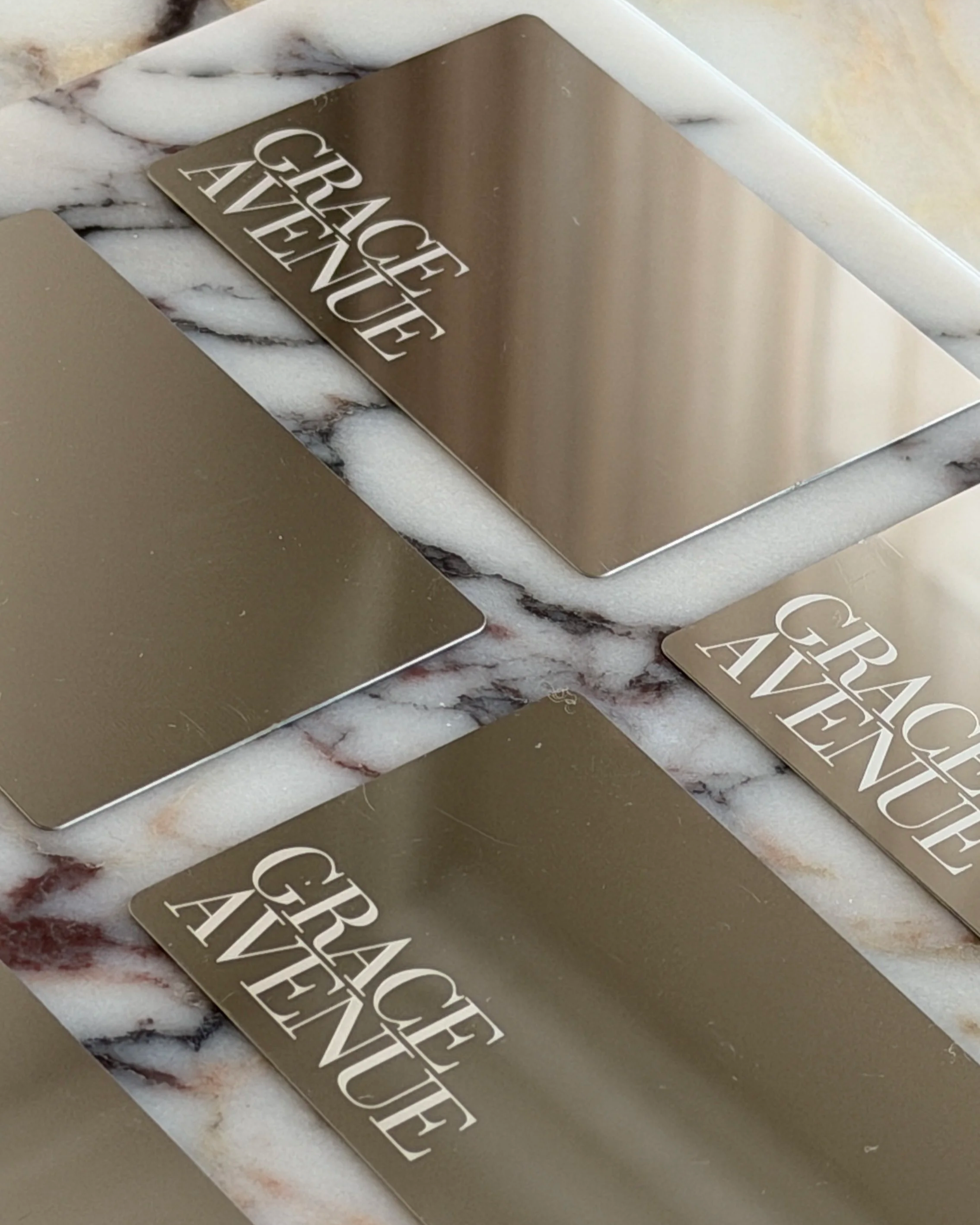 Close-up photo of a metallic business cards with engraved text that reads 'GRACE AVENUE'. The object is placed on a marble surface with a white and black veined pattern. Grace Avenue is the home to Shana Aesthetics and other beauty business owners.