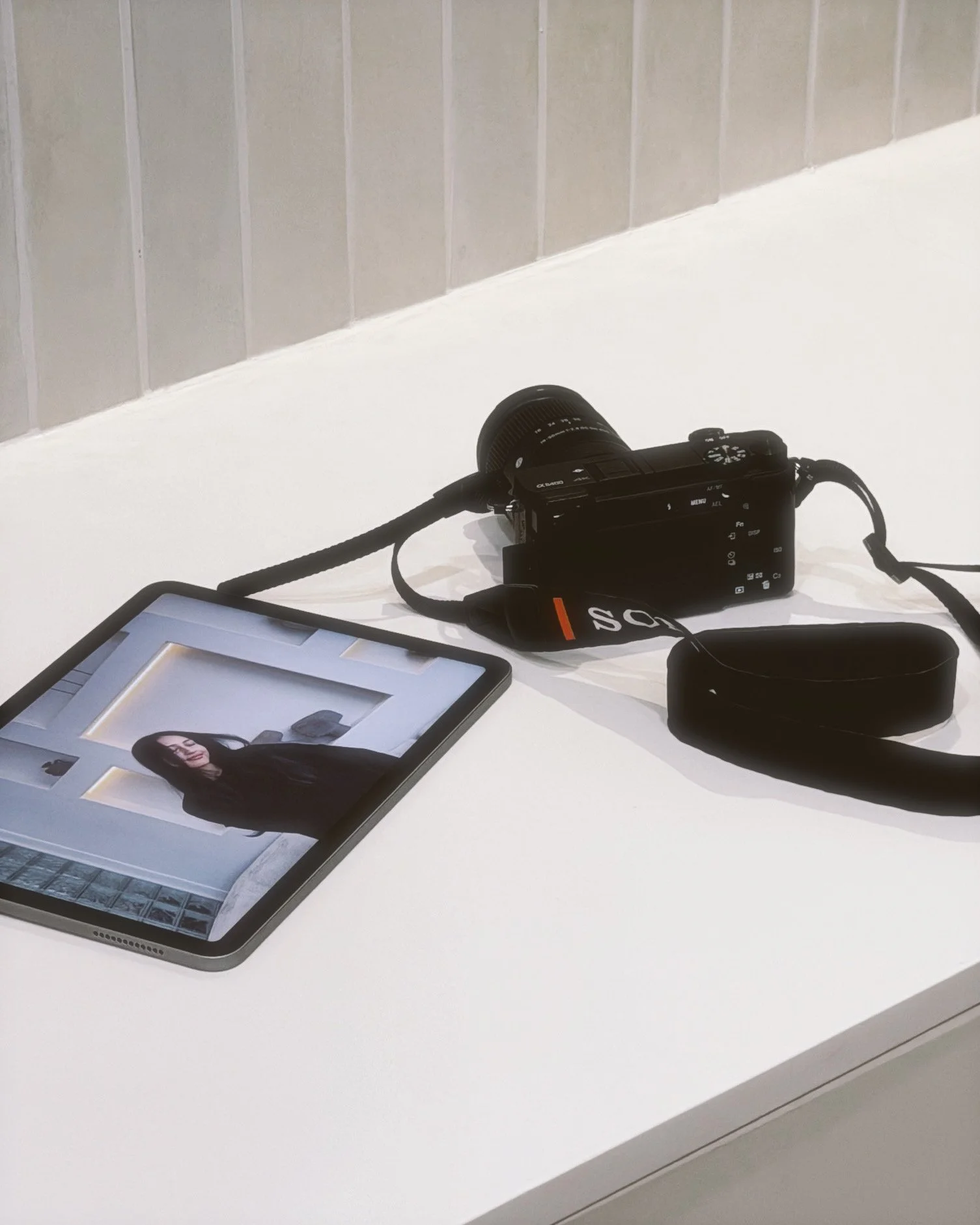Tablet displaying a portrait of a woman with black hair, a camera, and a black case on a white table against a plain wall