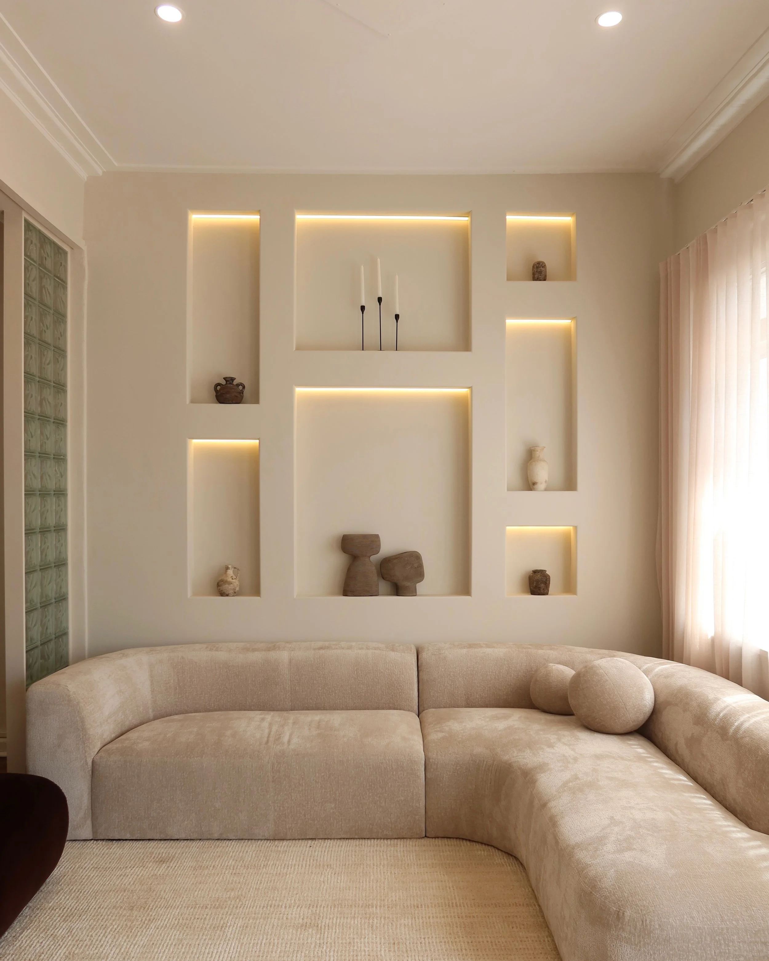 Living room with a beige sectional sofa and decorative pillows, a wall with built-in shelves displaying pottery and wooden sculptures, and a window with sheer curtains.