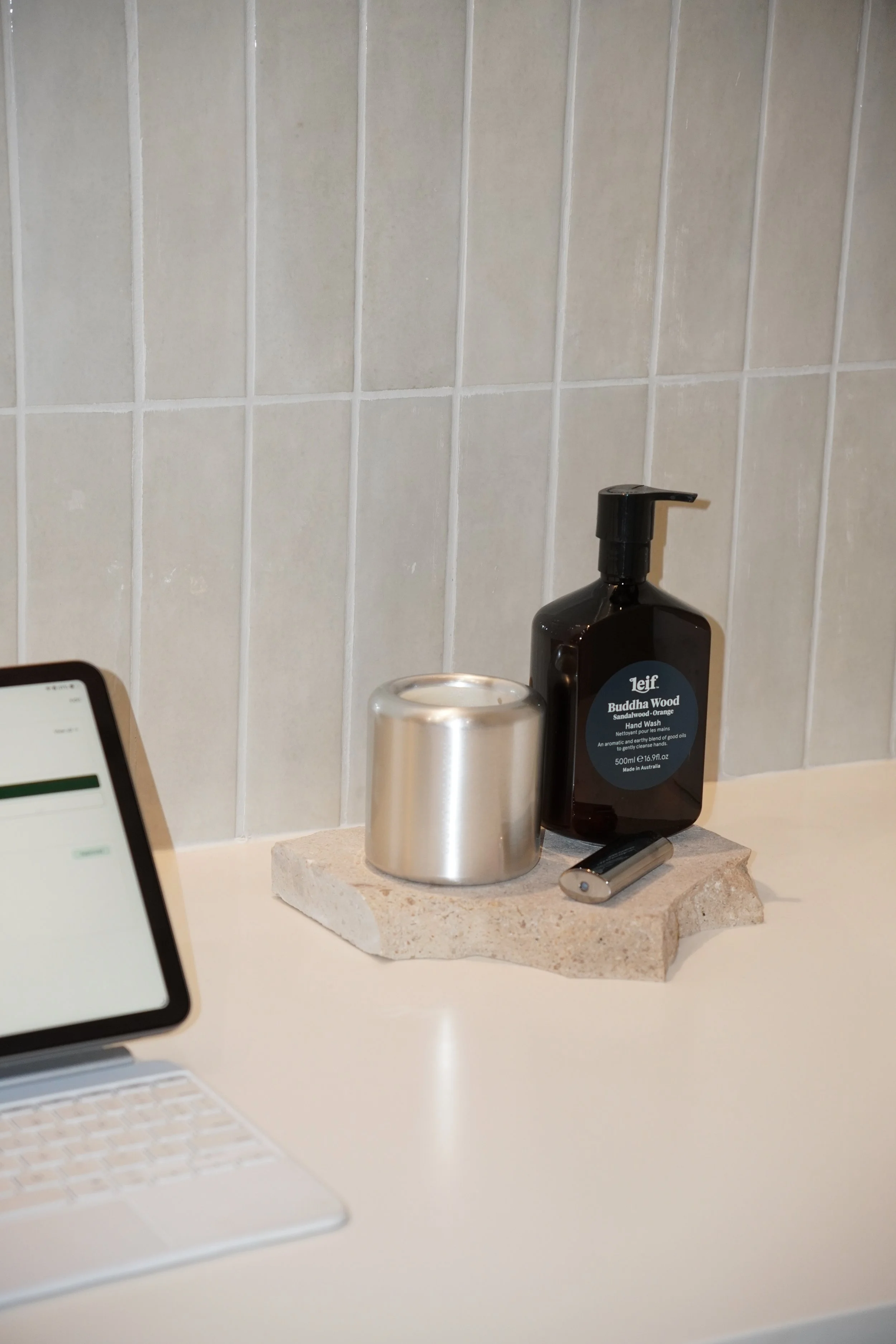 Bathroom countertop with a black hand soap dispenser labeled 'Leif Buddha Wood Sandalwood & Orange Hand Wash', a silver candle, a small cylindrical object, and part of a laptop and a phone visible in the foreground. The background features a beige ti