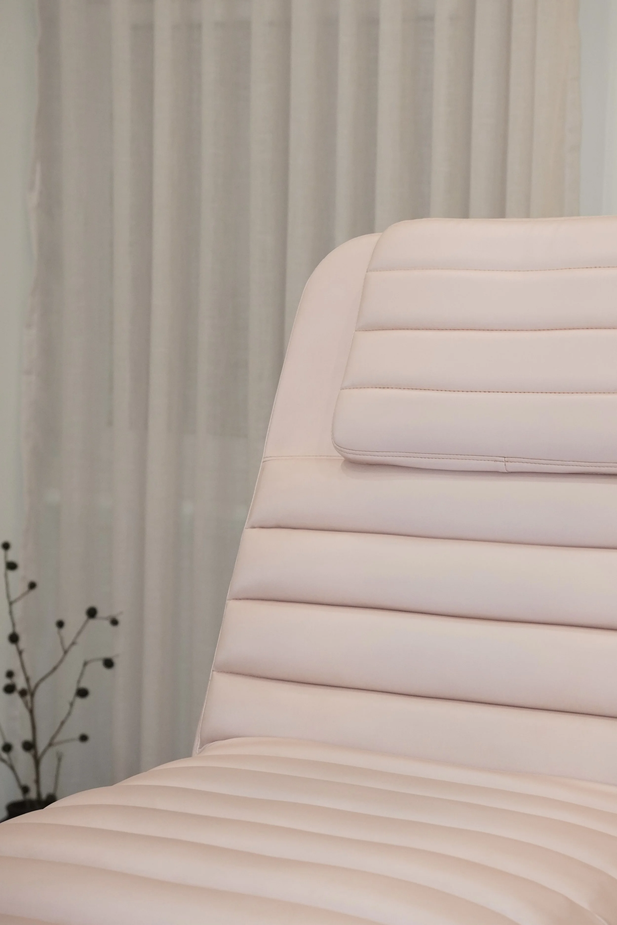 Close-up of a white, padded treatment chair with horizontal stitching, next to beige curtains and a small part of a decorative branch in the lower left corner. Shana Aesthetics treatment room. 