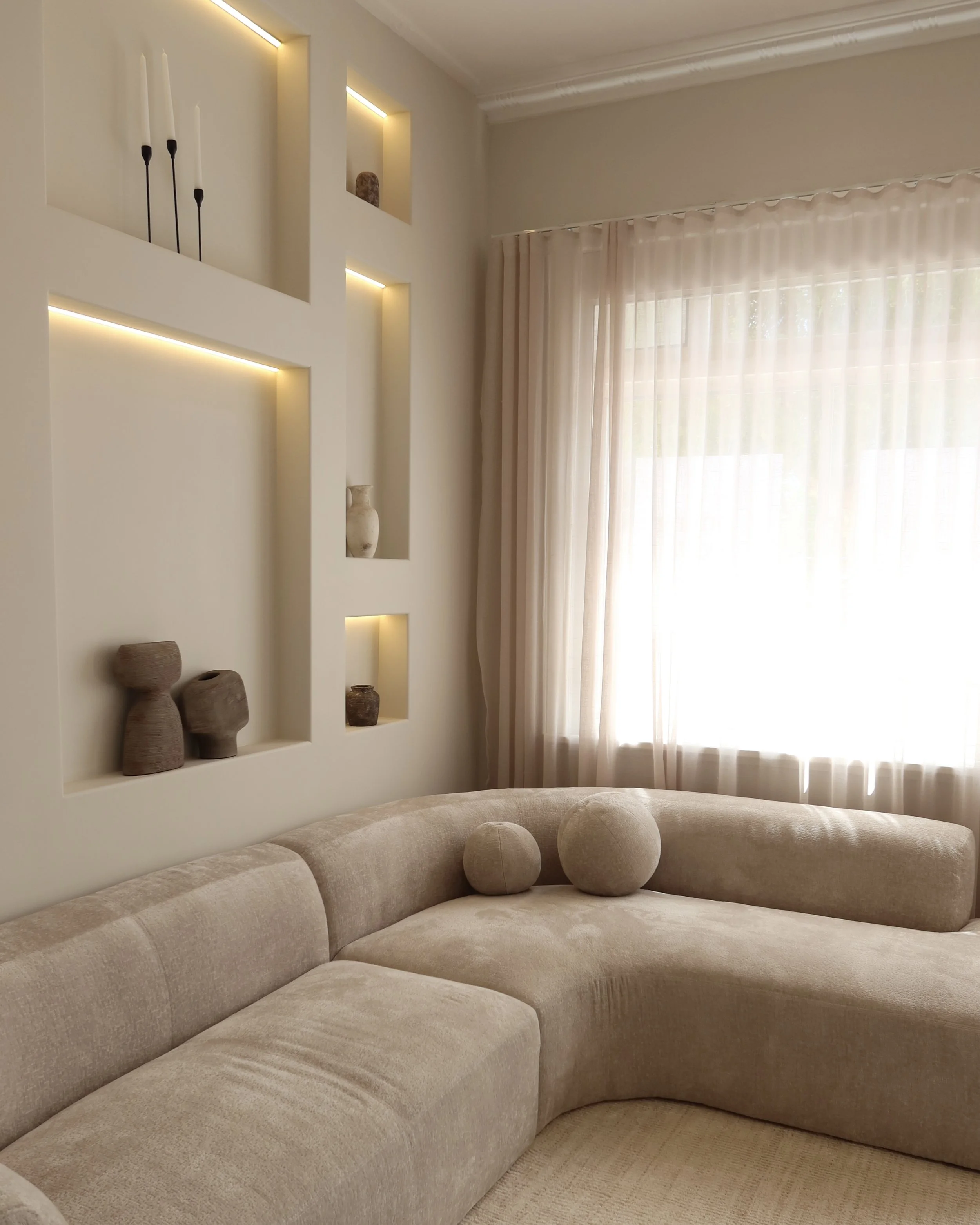 Living room with a beige curved sofa, decorative pillows, built-in wall shelves with vases, sheer curtains, and natural light.