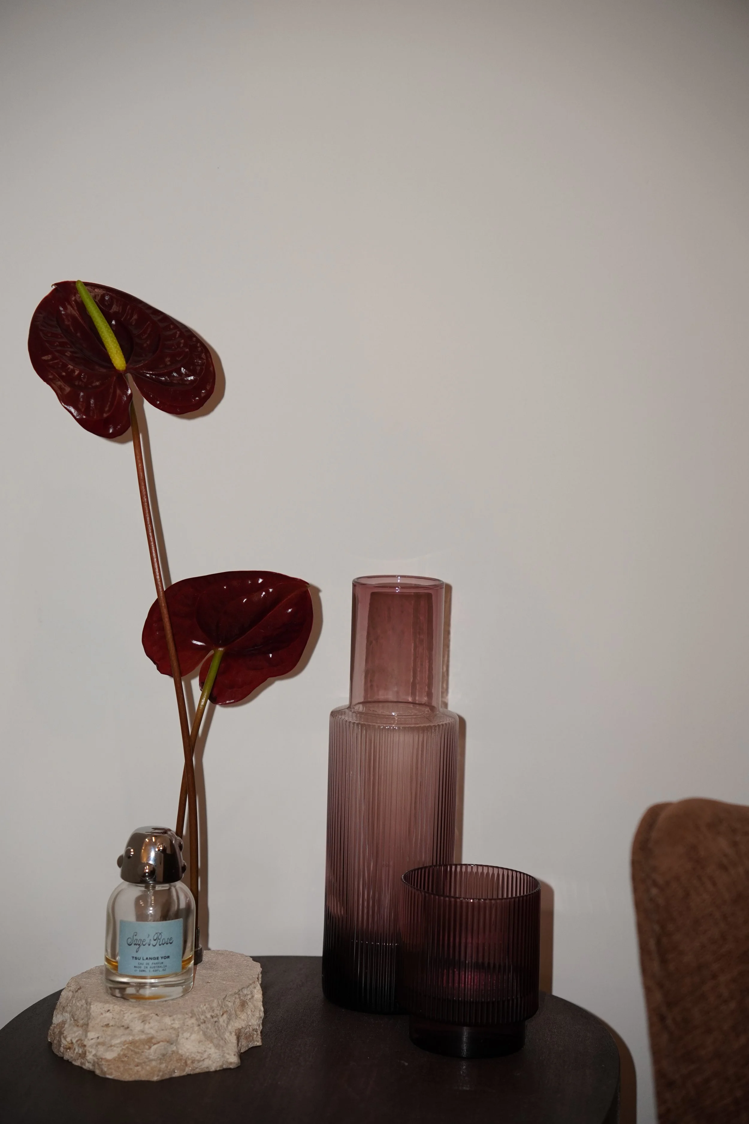 A decorative arrangement on a black round table featuring a pink ribbed glass vase, a matching pink ribbed glass container, a clear perfume bottle with a white label and metal spray top, and a soap dispenser. There are also two dark red anthurium flo