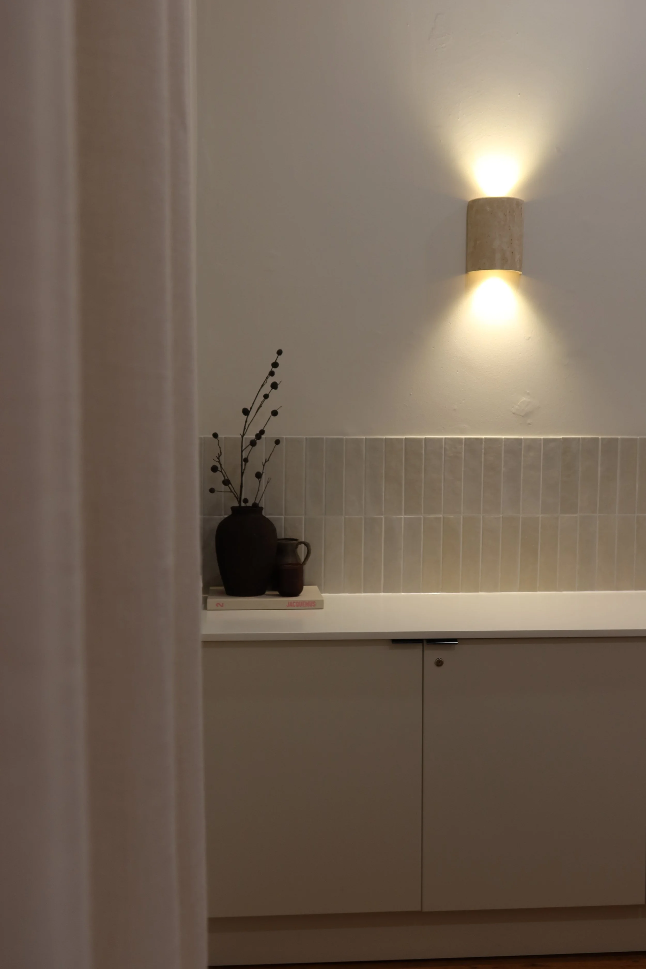 Partially concealed view of a minimalist interior with beige cabinetry, a white countertop, a tiled backsplash, a wall-mounted light fixture, and a decorative black vase with dried branches on a book.