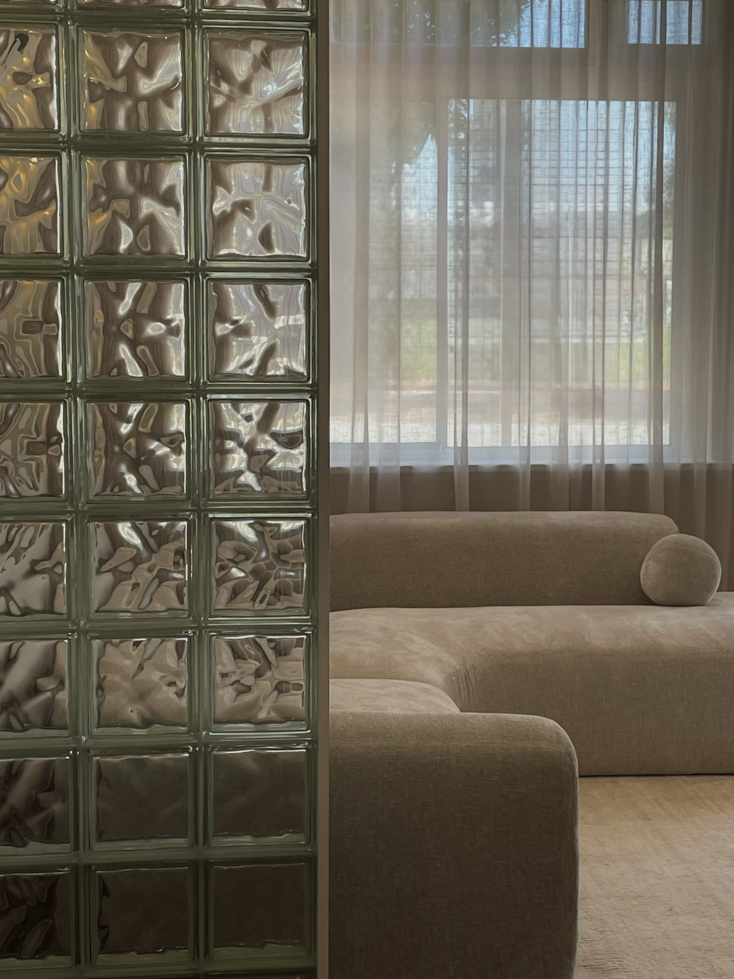 Interior view of a room with a beige sofa and a window with sheer curtains, partially divided by a glass block wall.