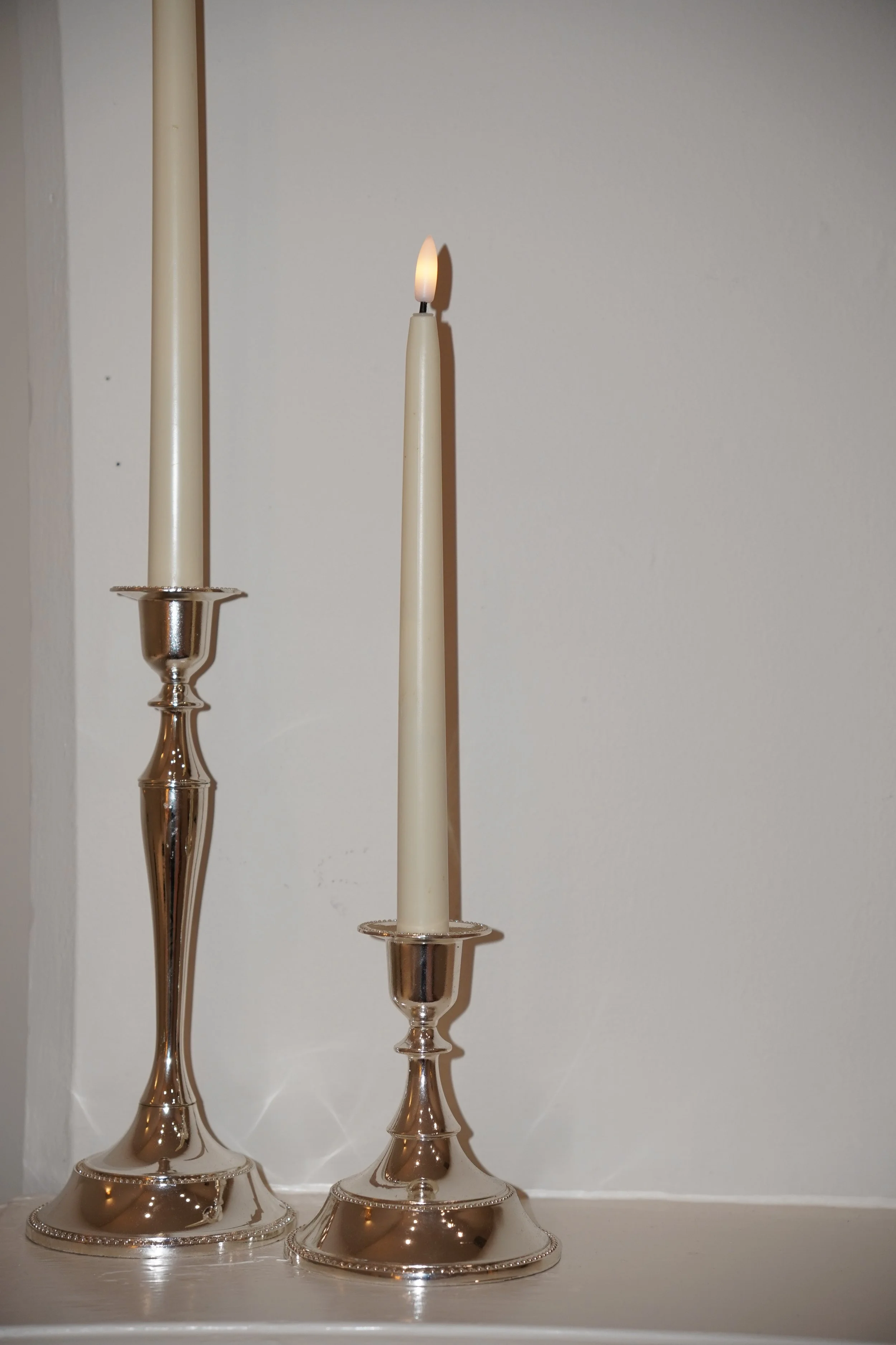 Three tall, elegant silver candlesticks with cream-colored candles, one of which is lit, placed on a white surface against a white wall.