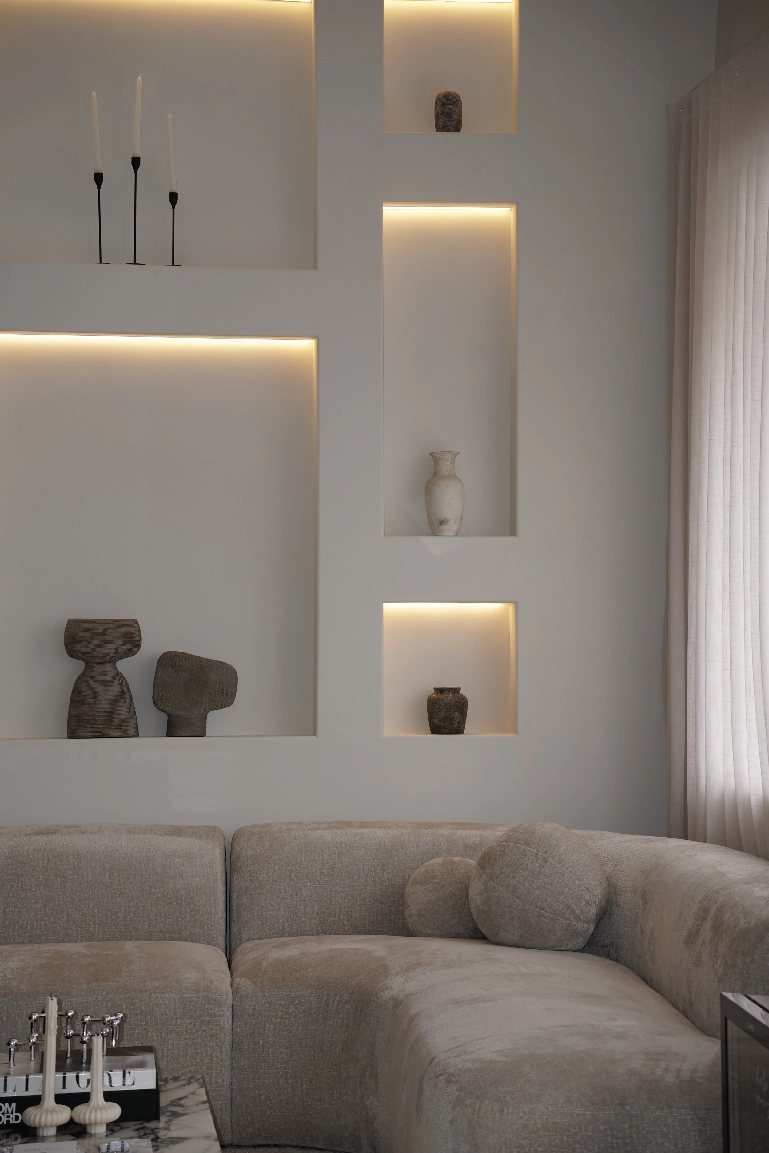 Modern living room with a beige sectional sofa, decorative vases on lit wall niches, and a window with sheer curtains.