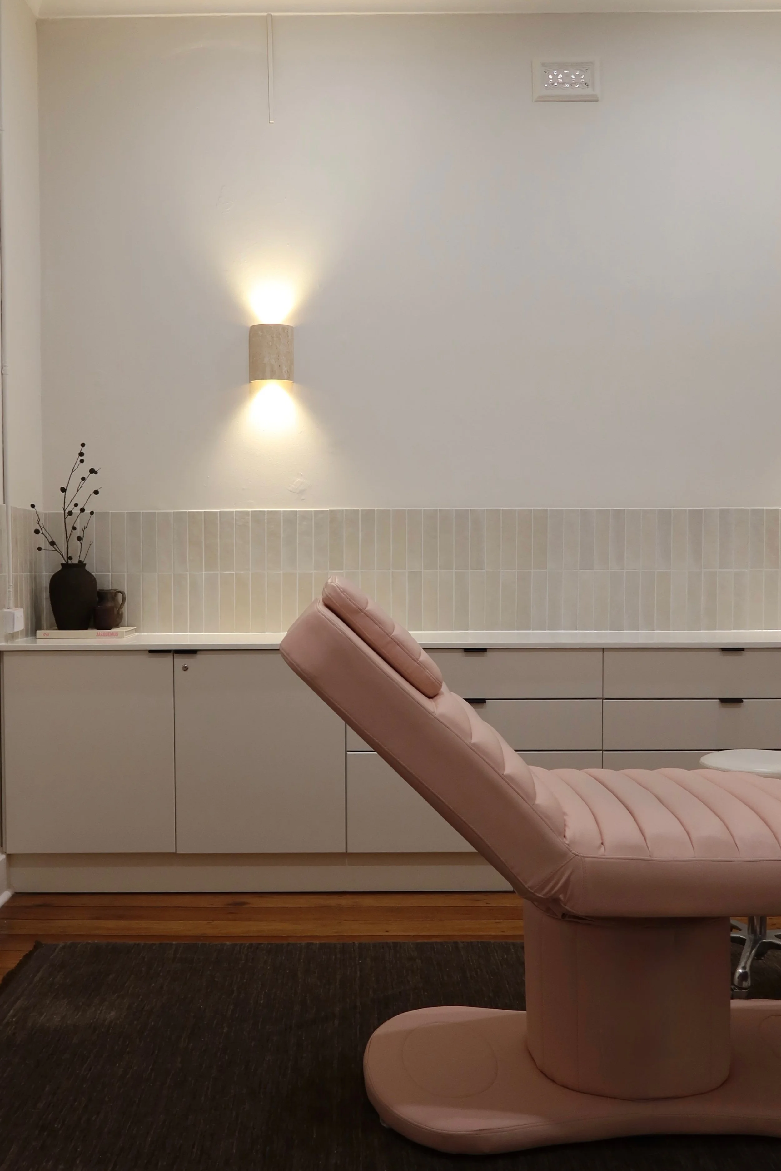 A medical or spa examination room with a pink adjustable chair, a white cabinet with drawers, a dark vase with decorative branches, a wall-mounted light fixture, and a dark rug on a wooden floor.