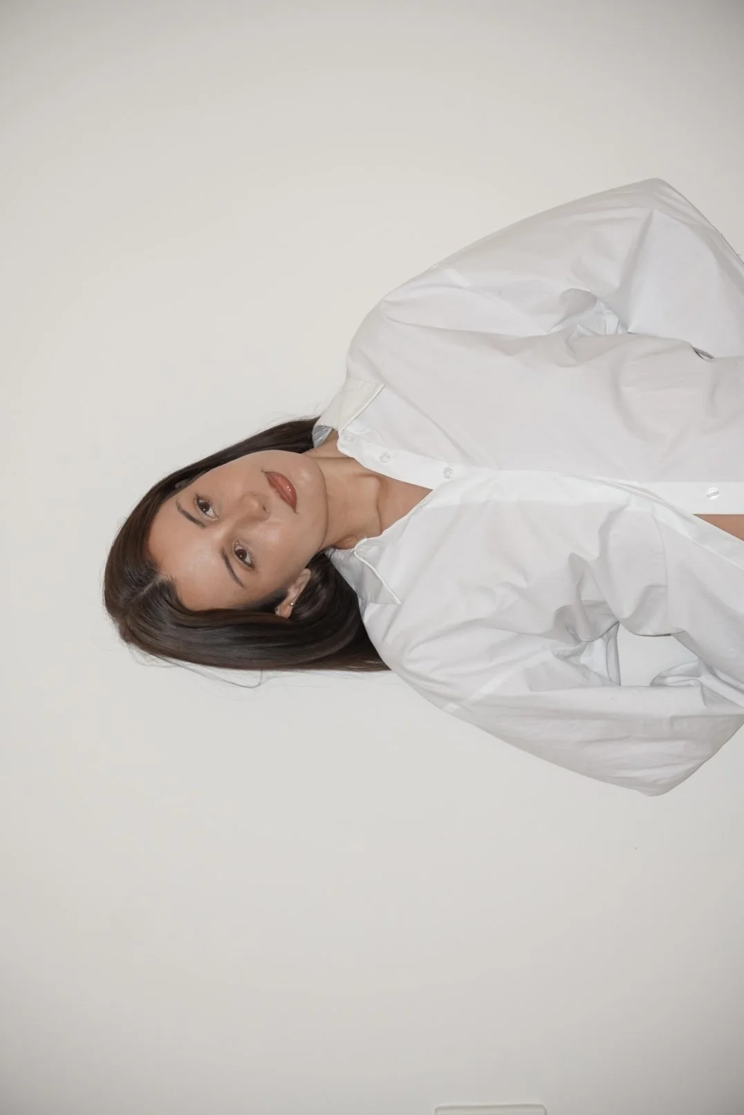 A woman with shoulder-length dark hair and a white button-up shirt, lying down against a plain white background, facing the camera. She's a Perth Cosmetic Nurse and does botox, filler, facial balancing, cheek filler, lip filler. 