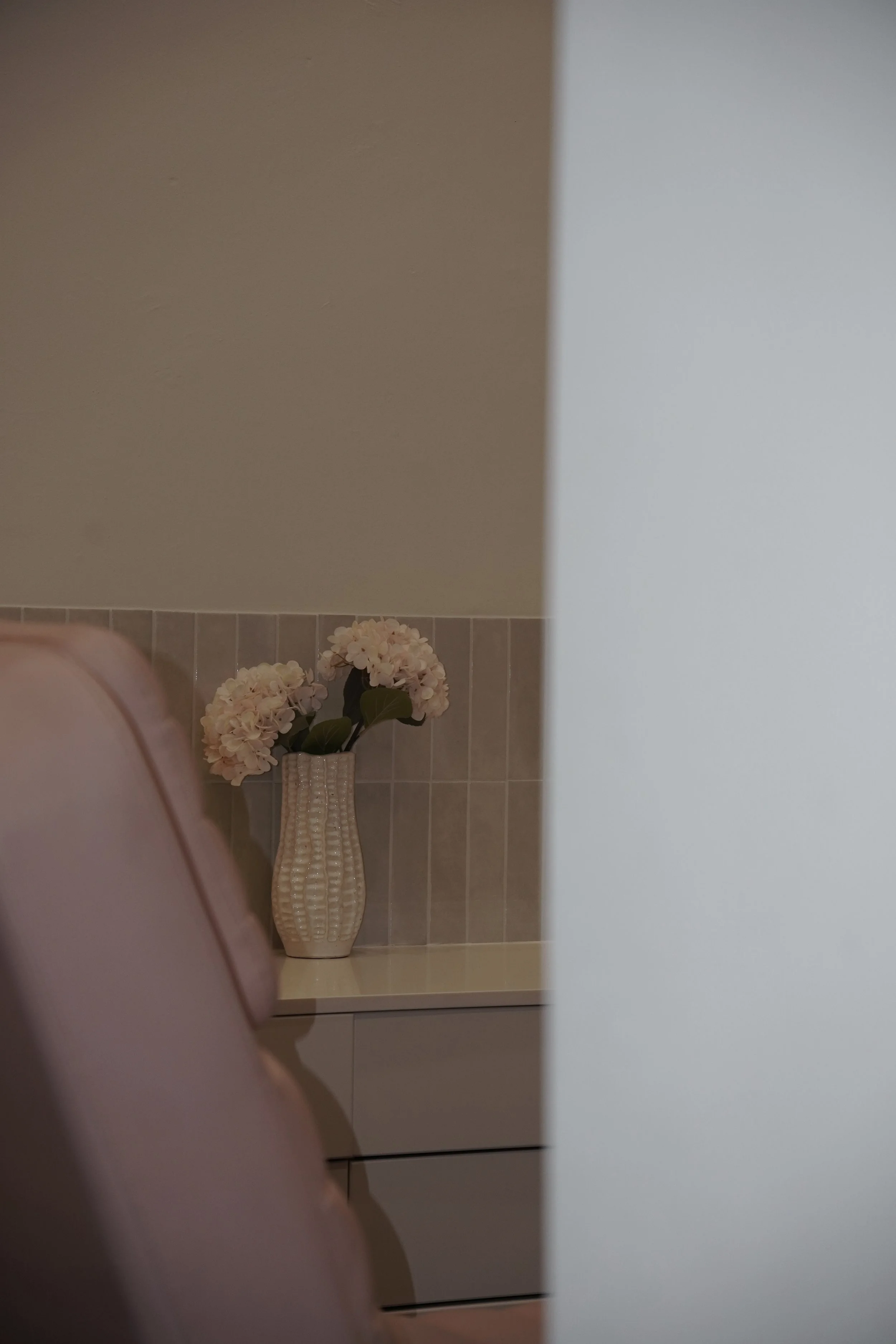 A view of a bathroom corner with beige and gray tiled walls, a tall beige vase with a textured pattern, and pink and white artificial flowers inside the vase.