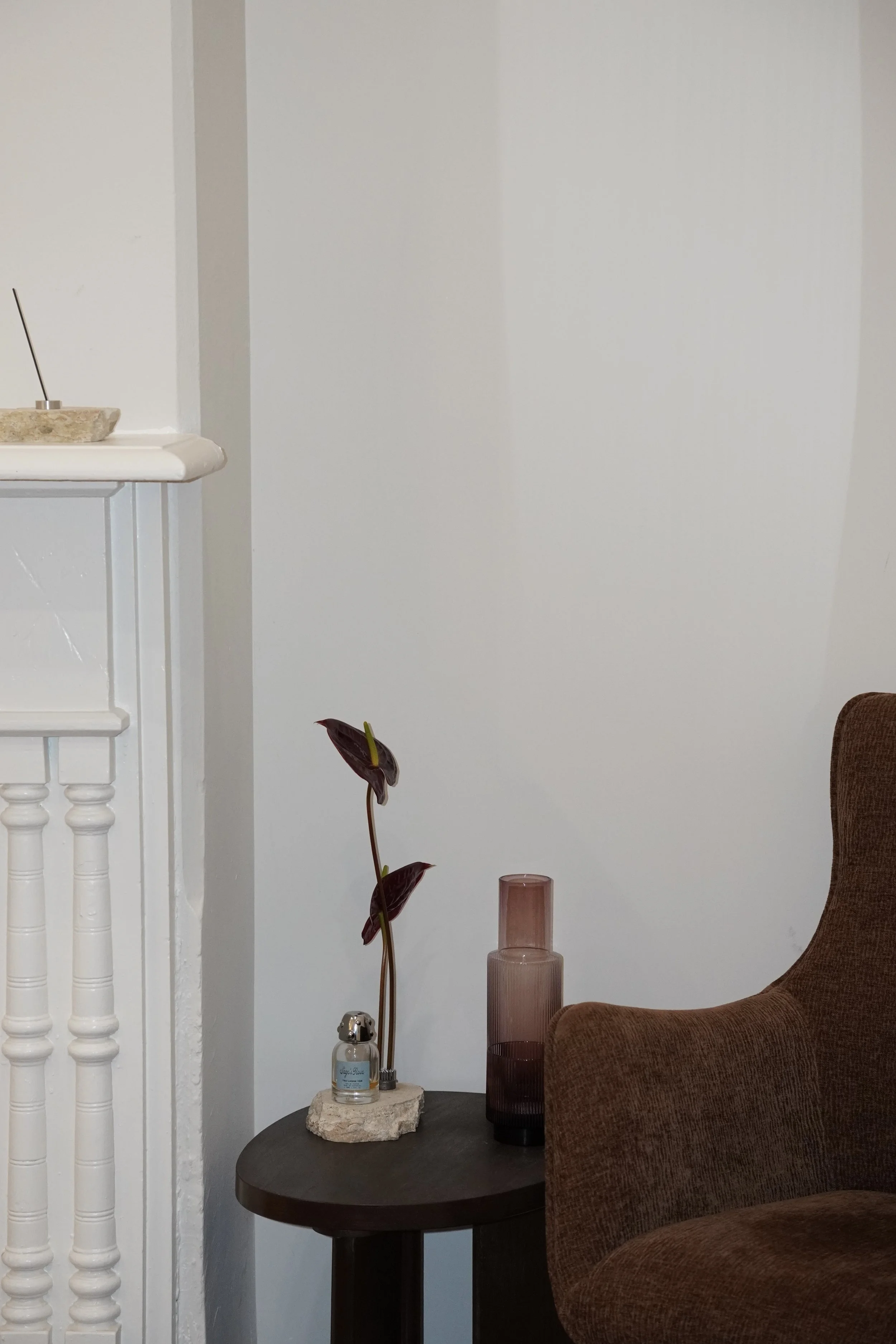 A living room corner with a brown upholstered chair, a dark round side table, and decorative items including a tall pink glass vase, a spray bottle, a small bottle on a stone slab, and a potted plant with dark red leaves. There is a white wall backgr