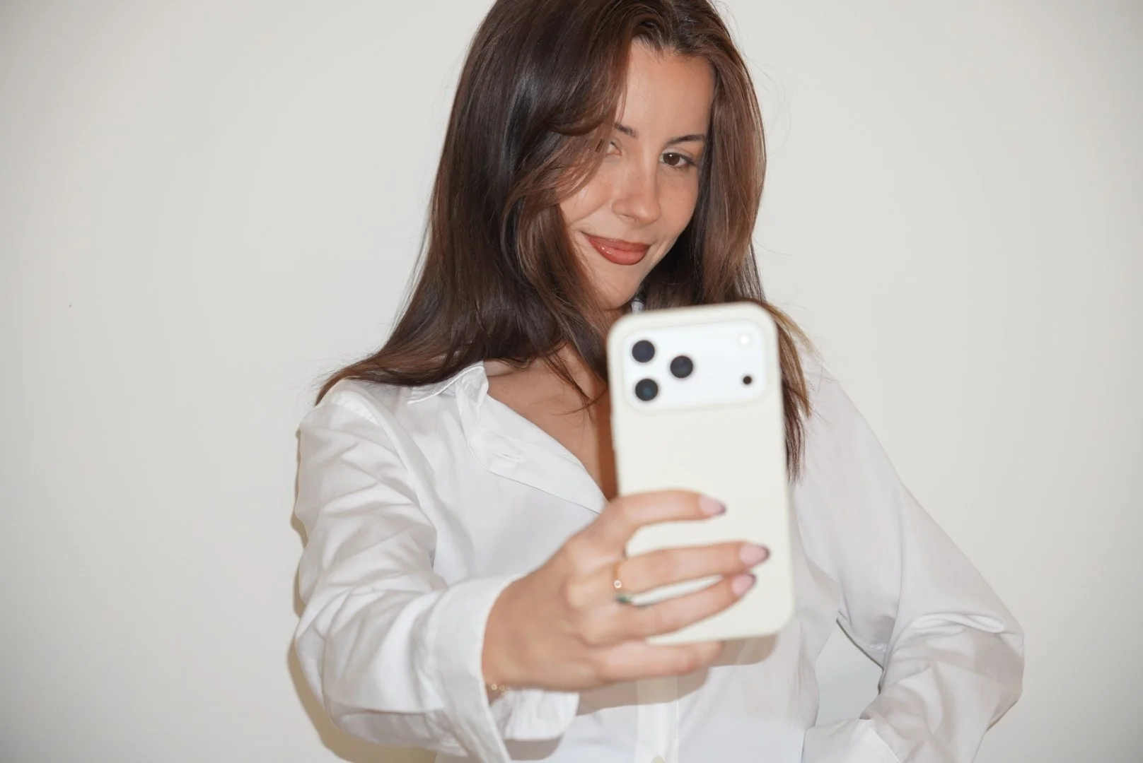 A woman with brown hair and wearing a white shirt taking a selfie with a smartphone