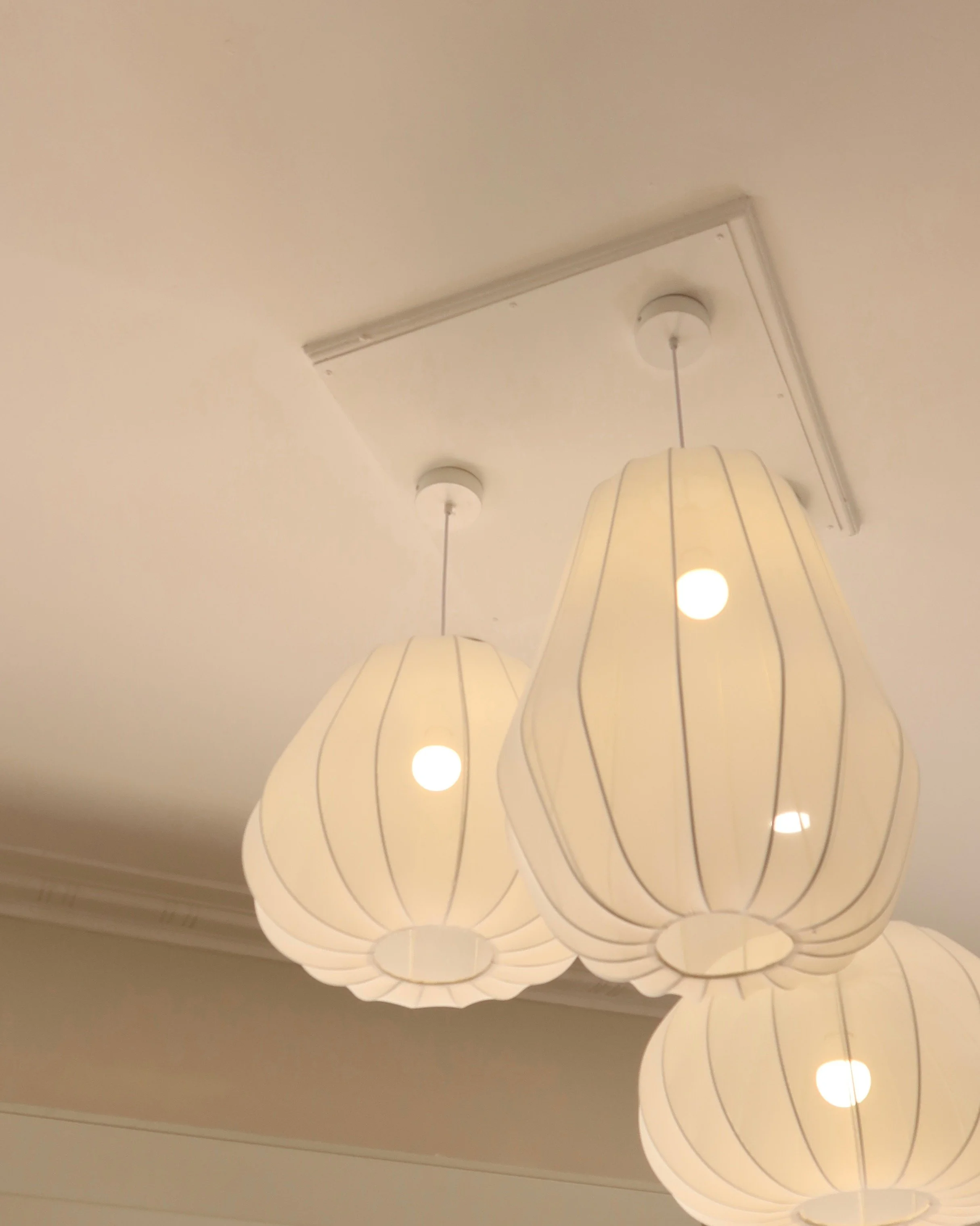 Three hanging pendant lights with white, fabric shades and internal light bulbs, mounted on a ceiling with a rectangular ceiling plate.