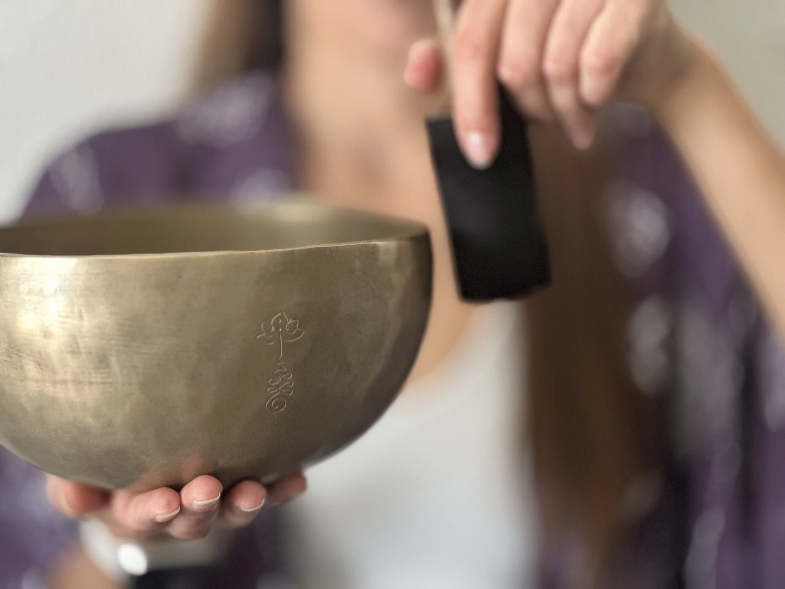 Close-up of hands holding a bronze singing bowl while striking it with a black mallet, capturing a moment of sound healing practice.