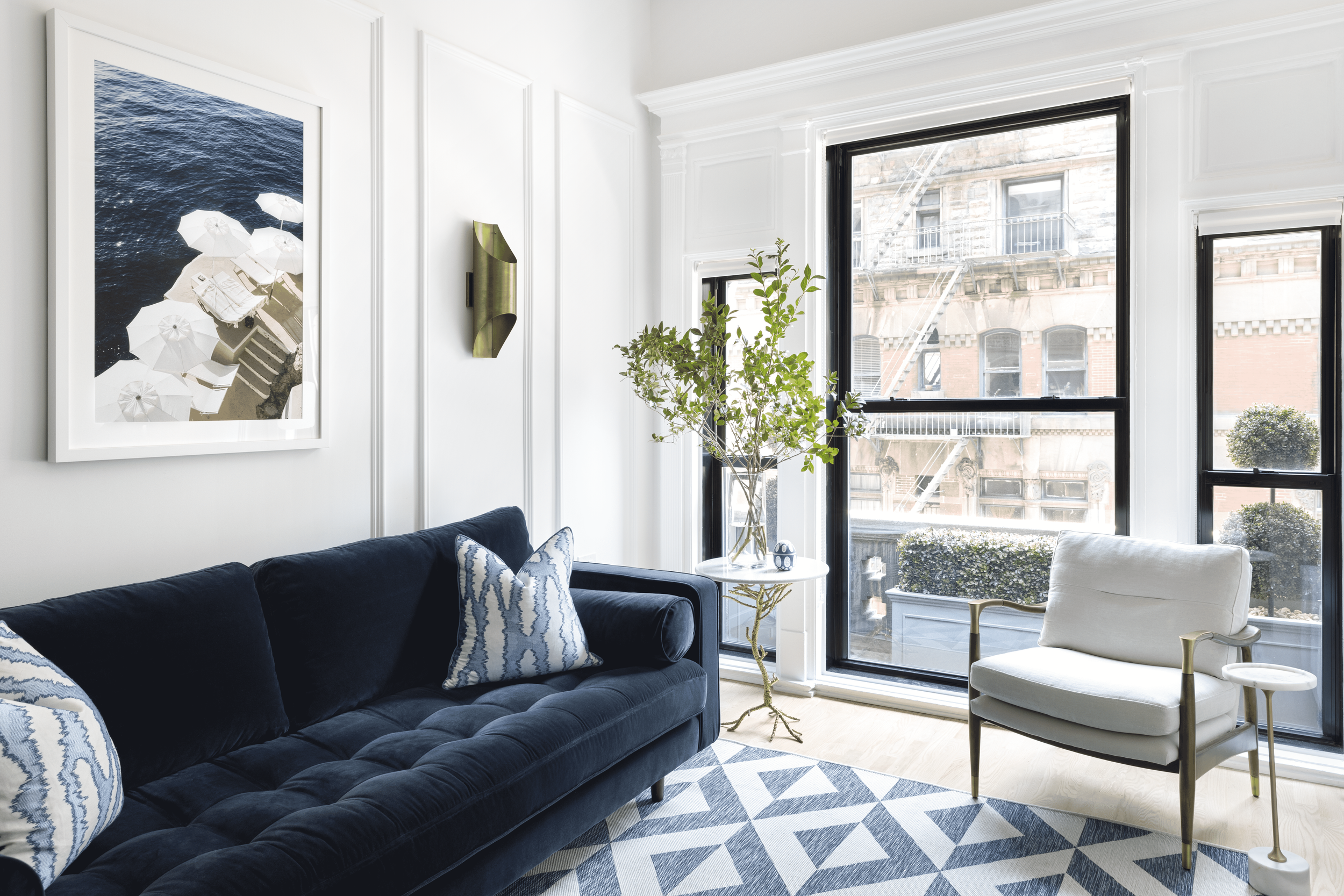 Living room with white walls, large windows showing city buildings, dark blue sofa with patterned pillows, white armchair, glass side table with a plant, patterned area rug.