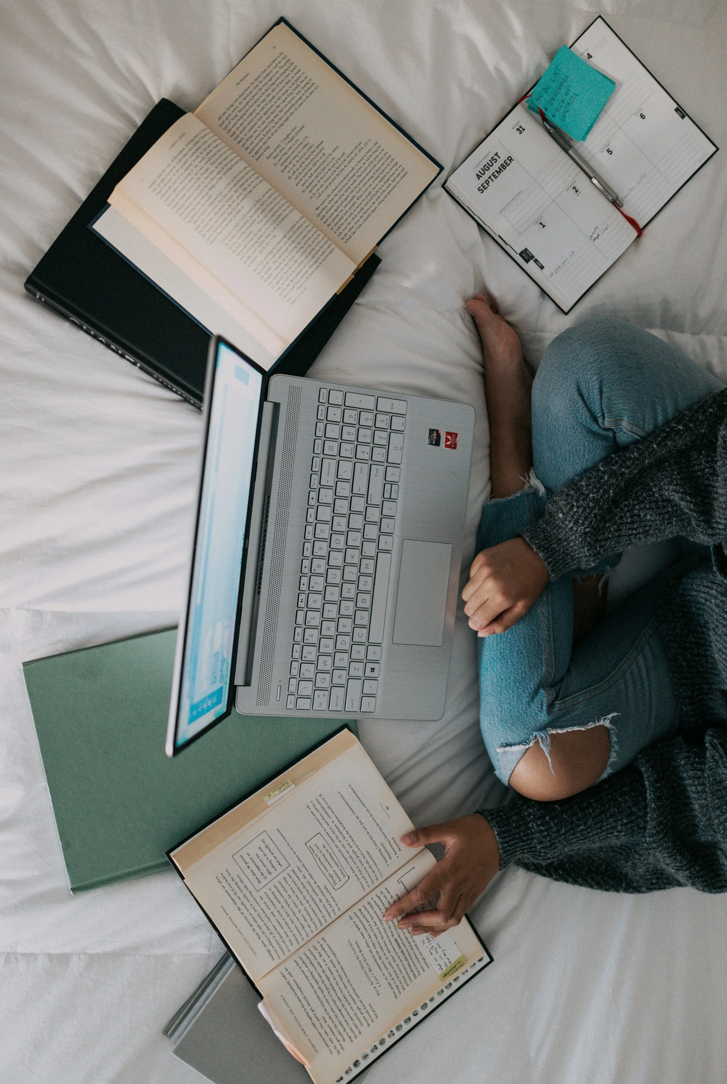 Overhead view of a person sitting on a bed surrounded by open books, a laptop, and a planner with sticky notes, working or studying.
