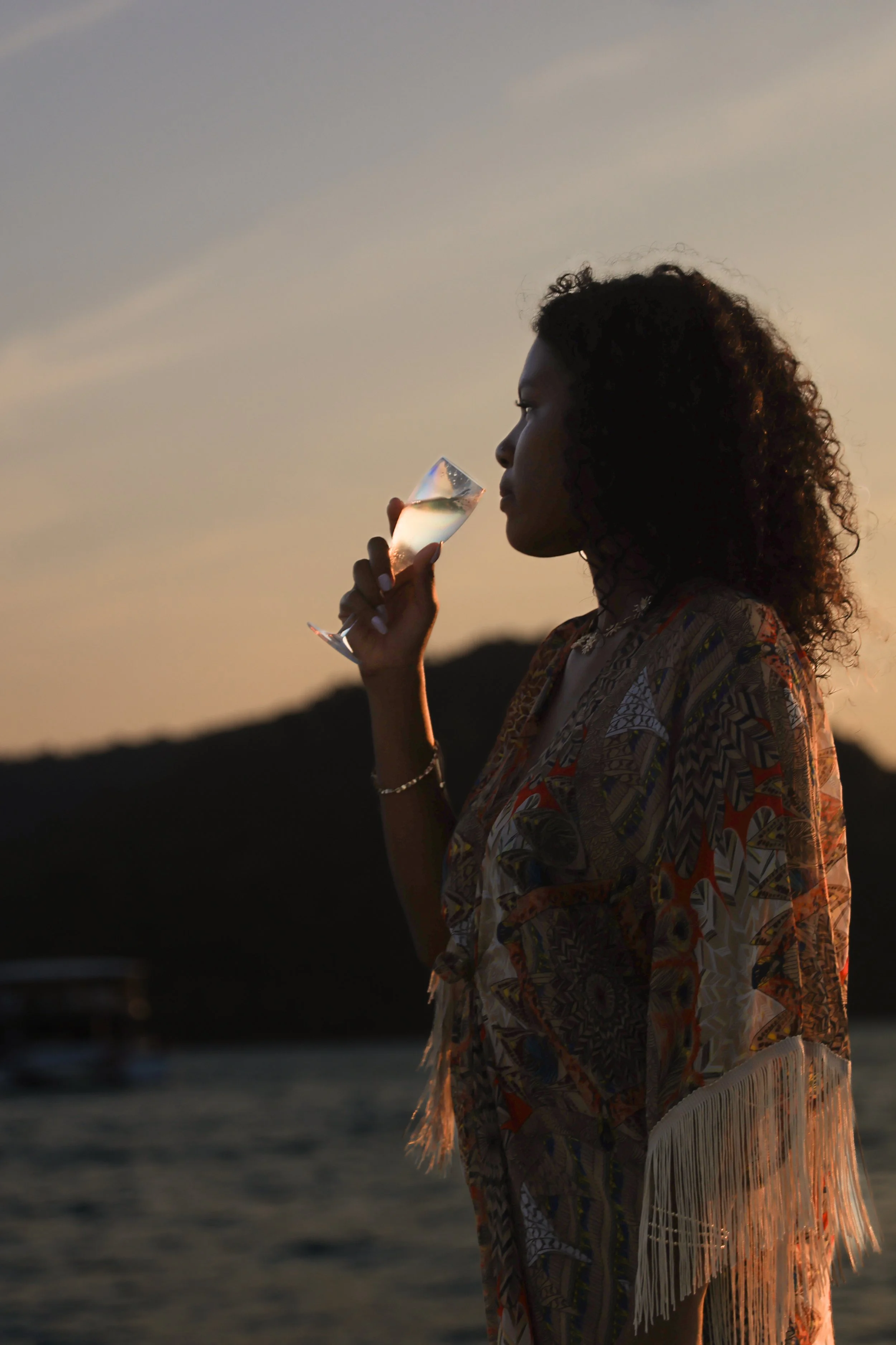 A woman with curly hair holding a glass of champagne while standing outdoors at sunset near water, with a mountain in the background.