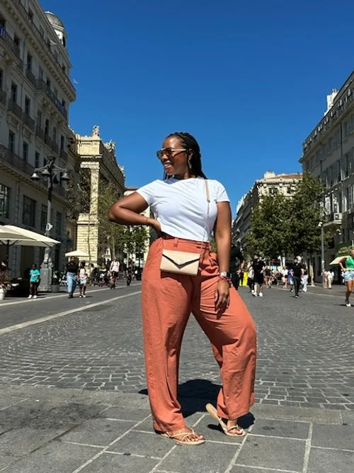 A woman standing in the middle of a busy city street, smiling and looking to her right, wearing sunglasses, a white t-shirt, peach high-waisted wide-leg pants, beige sandals, and carrying a beige purse, with historic buildings and other pedestrians in the background under a clear blue sky.