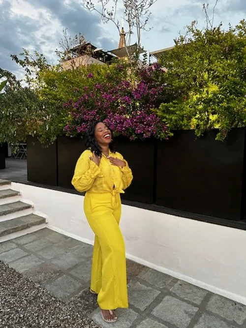 A woman in a bright yellow outfit standing outside near a white wall, surrounded by lush green plants and purple flowers, smiling and laughing.