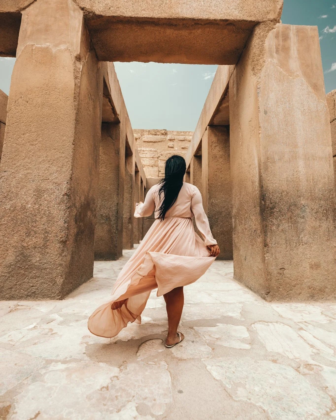 A woman in a flowing, light-colored dress walking through ancient stone ruins with large rectangular stone blocks and a clear sky overhead.