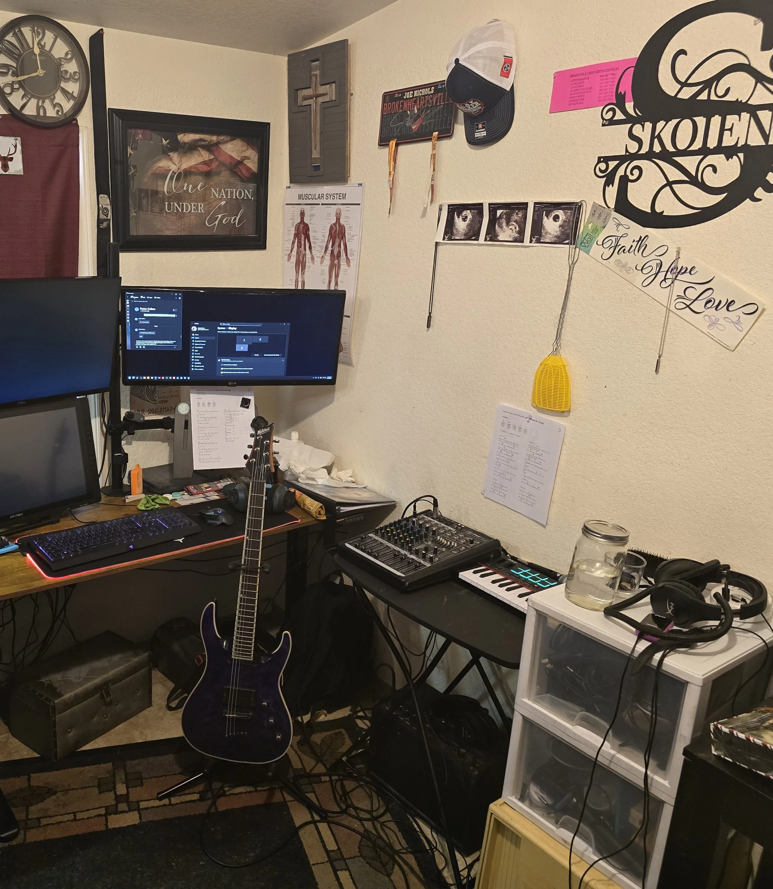 A music studio or workspace with a black guitar, computer monitors, a mixing console, and various posters and decorations on the wall. The wall displays a sign that says 'One Nation, Under God' and pictures of an eye. There are personal items like hats, a yellow plastic speaker, and a mason jar on a white plastic storage unit.