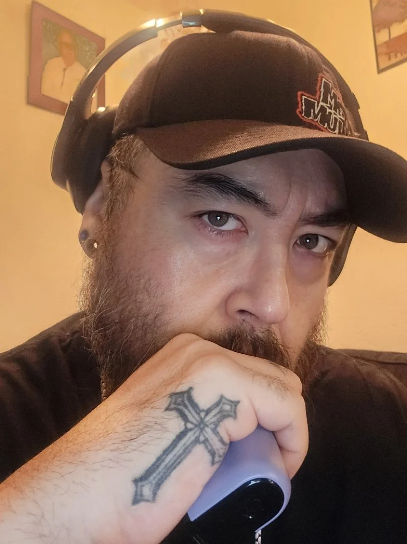 A man with a beard and short hair wearing a black hat and headphones, resting his chin on his hand with a tattooed cross on his knuckles, in a room with yellow walls and framed pictures.
