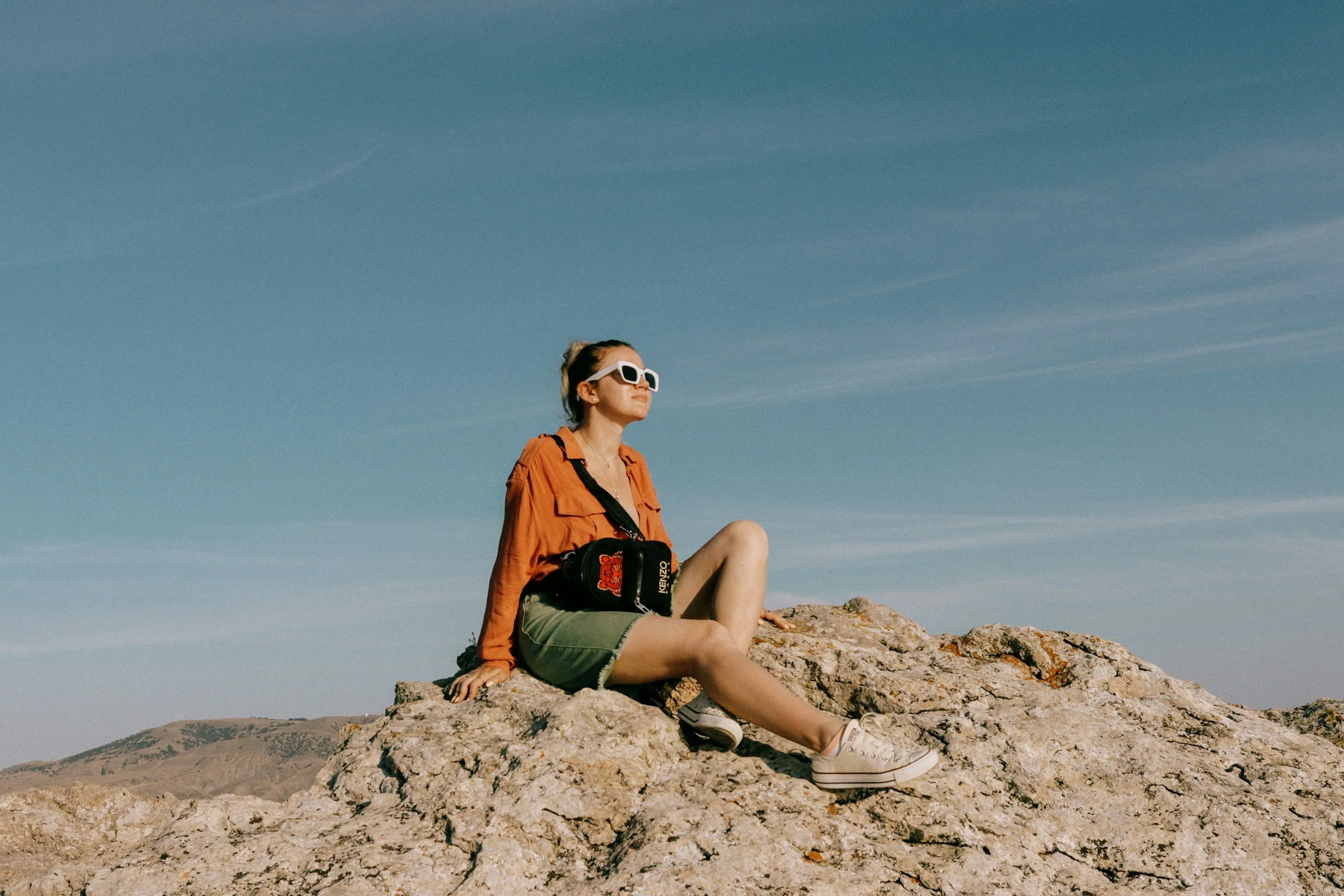 A woman sitting on a large rock formation outdoors, wearing white sunglasses, an orange jacket, green shorts, and white sneakers, with a black bag across her shoulder, under a vast blue sky with minimal clouds.