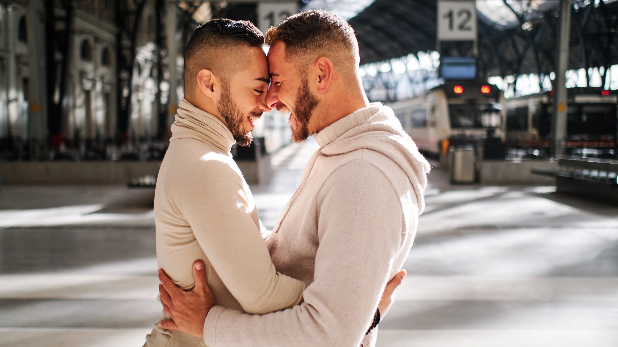 Two men embrace in a tender moment at a train station, smiling with foreheads touching, surrounded by train tracks and platforms.