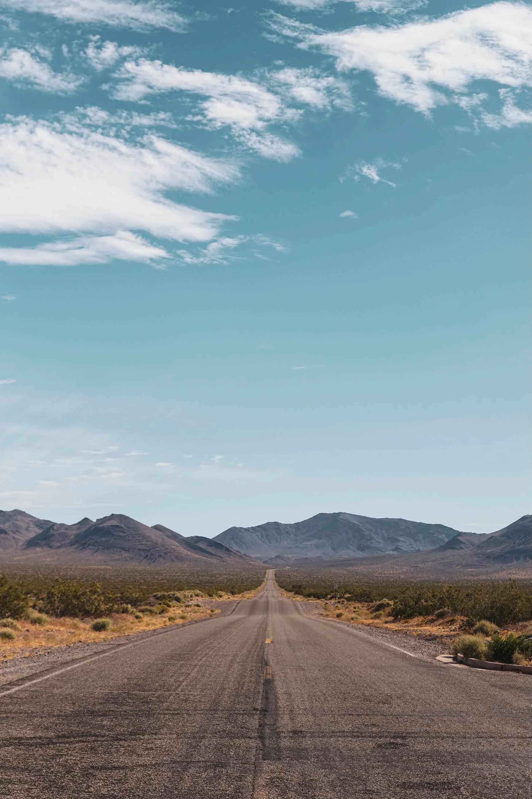 A long, straight desert highway stretching toward distant mountains under a partly cloudy sky.