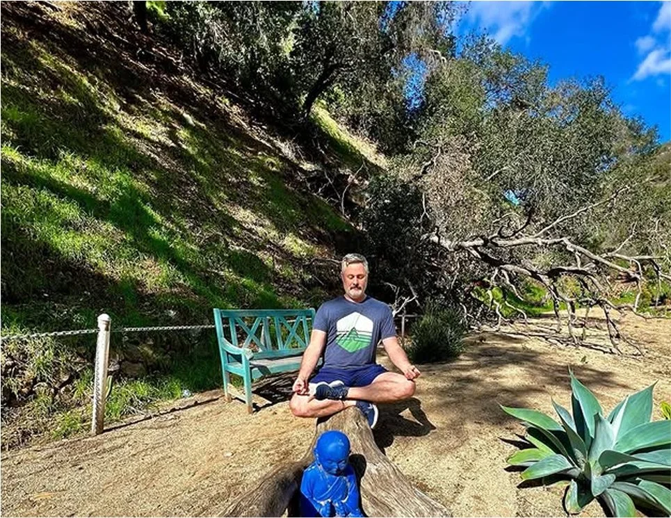 A man sitting cross-legged on a fallen tree in a park, meditating with eyes closed. He is wearing a navy t-shirt and shorts. There is a blue statue of a meditating figure in front of him, and a large tree with sprawling branches in the background.