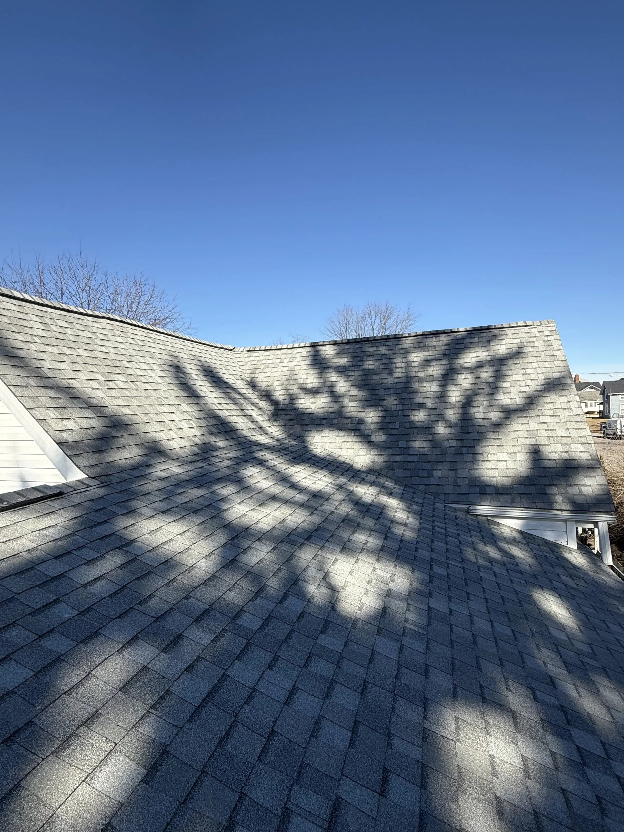 Shaded roof with gray shingles on a house under a clear blue sky, with tree shadows cast on the roof. Roofing services in Lafayette, IN.