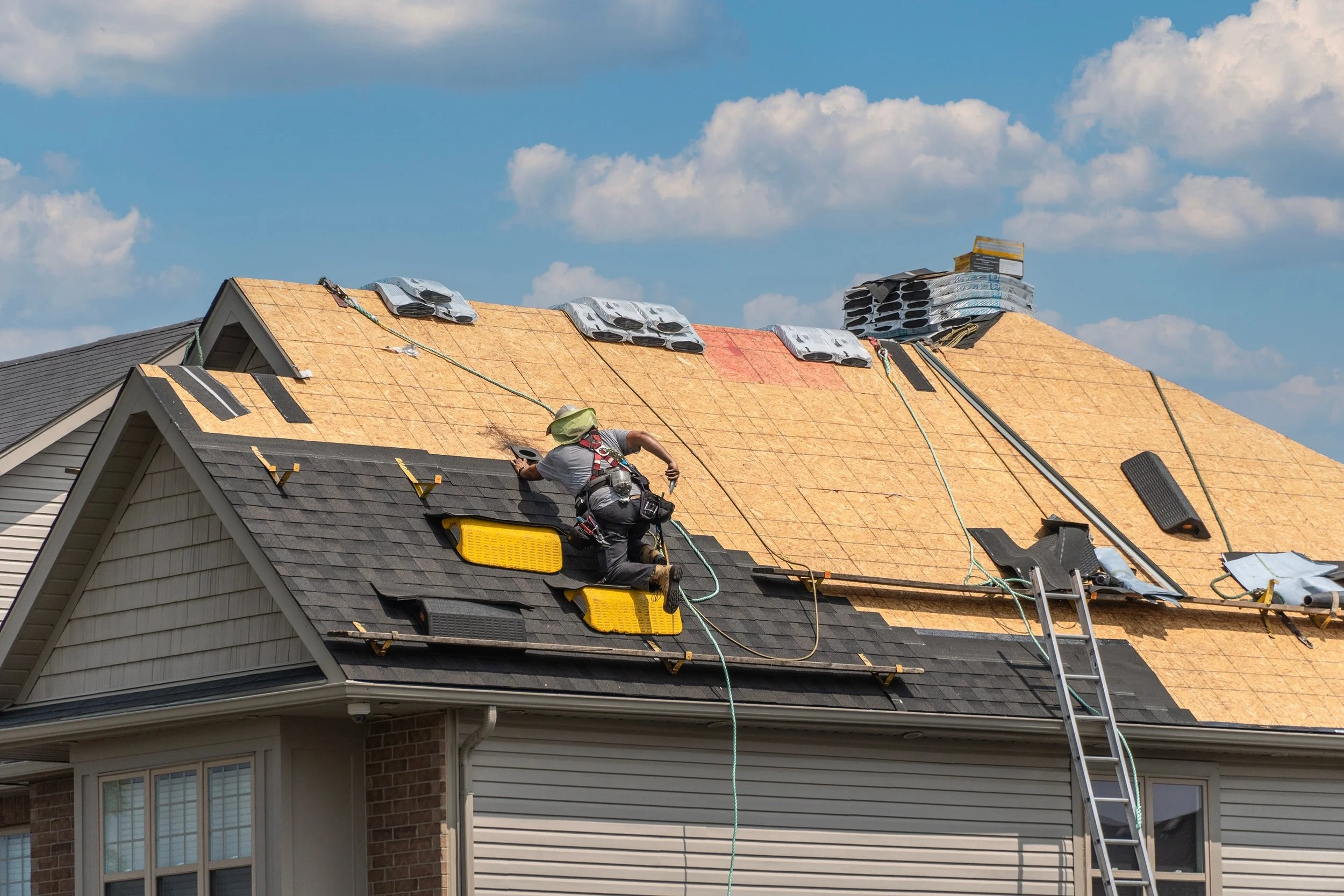 A construction worker on a roof installing new shingles, with some shingles and roofing materials scattered around, and a ladder leaning against the house under a blue sky with clouds. Roof replacement in Rossville, IN.