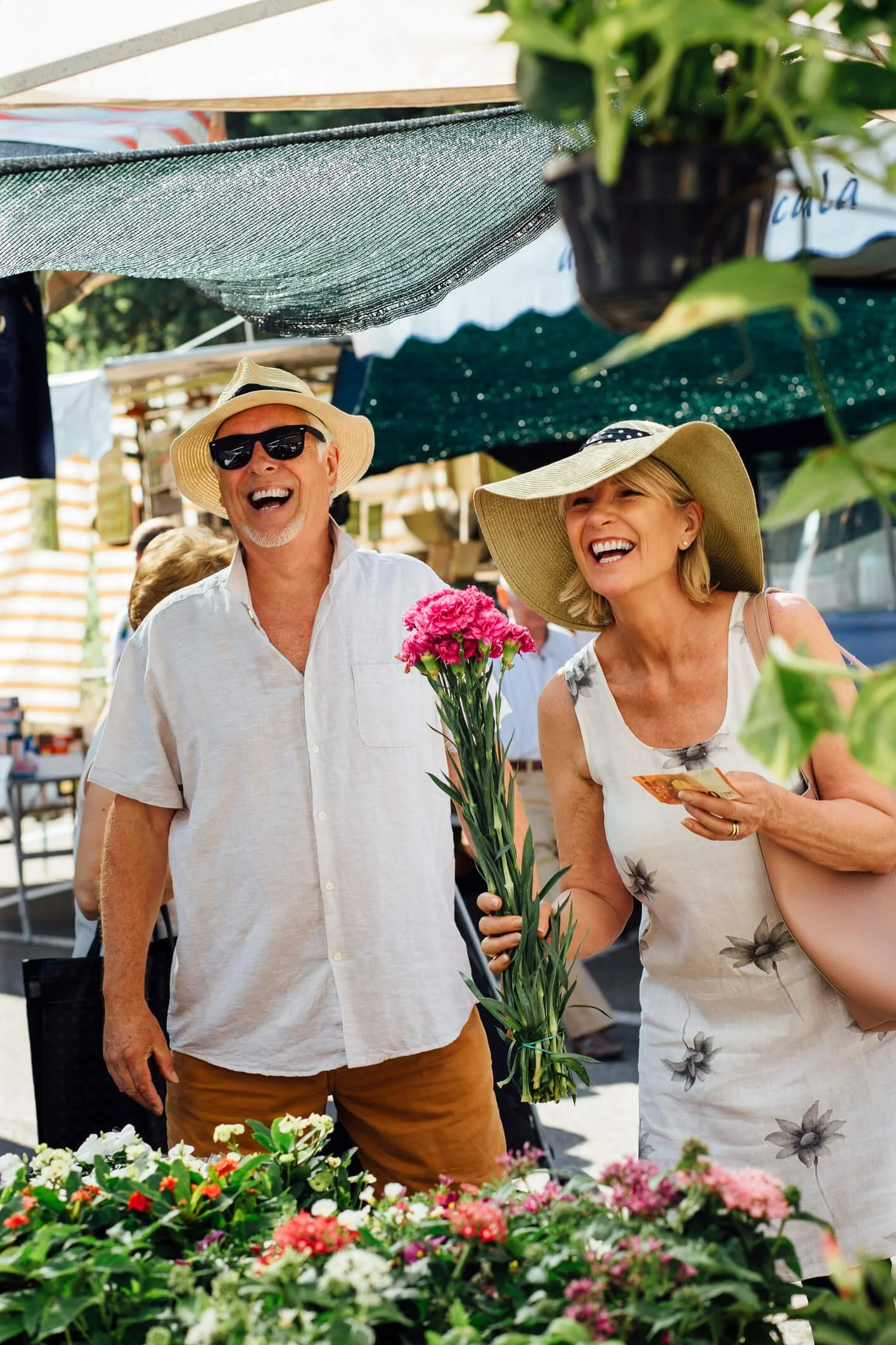 A smiling man and woman with wide-brimmed hats at an outdoor market, holding pink flowers and a brochure, with flower stalls in the background.