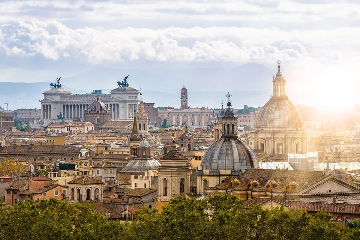 Cityscape of Rome, Italy, with historic domes, churches, and rooftops, under a partly cloudy sky with sunlight.
