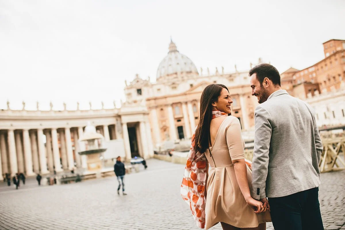 A smiling couple holding hands in front of St. Peter's Basilica in Vatican City.