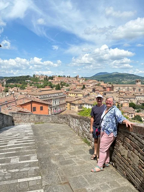Two women standing on a stone pathway overlooking a historic Italian town with terracotta roofs and rolling hills under a partly cloudy sky.