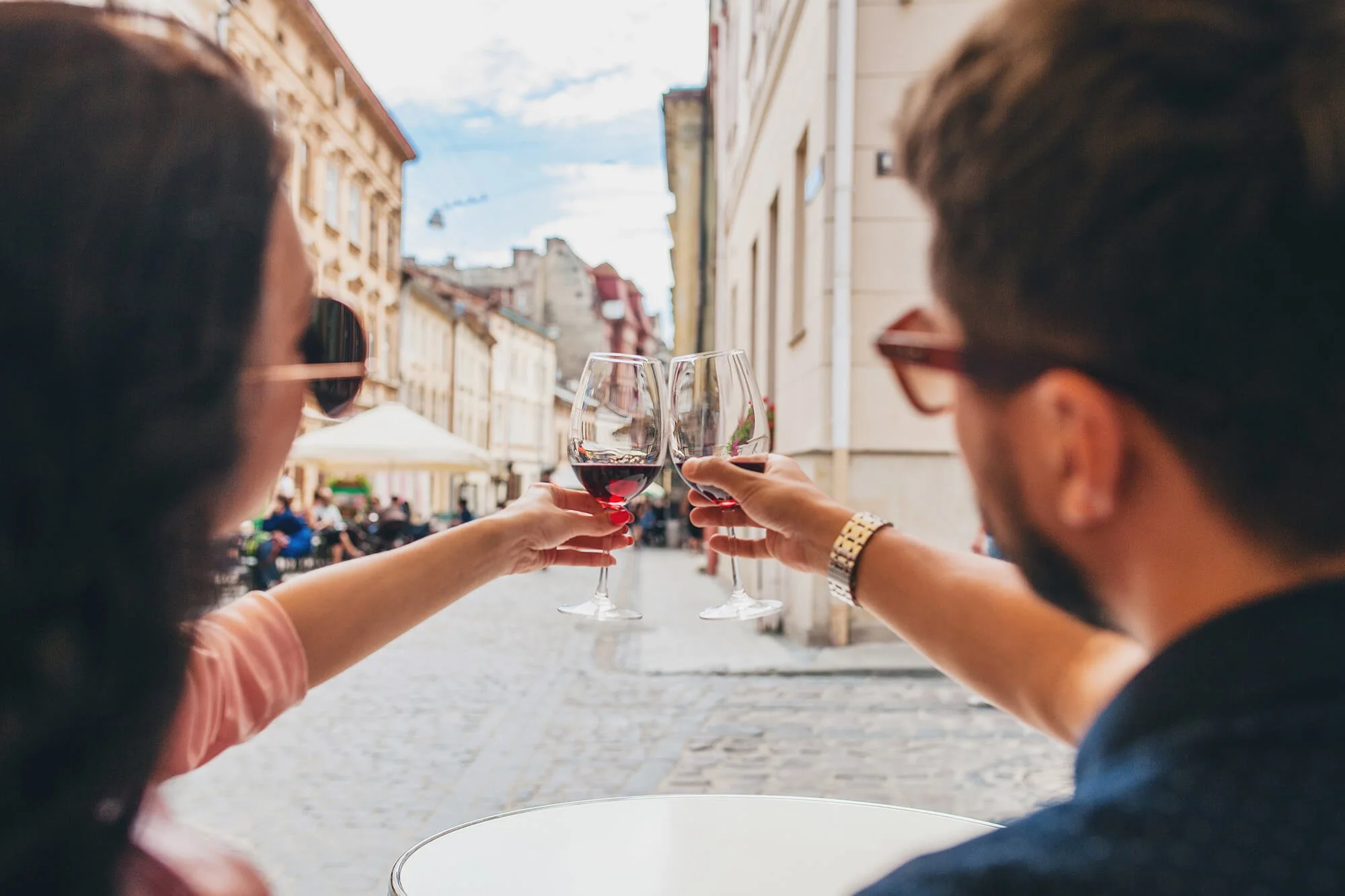 Two people clinking glasses of red wine outdoors on a cobblestone street in a city.