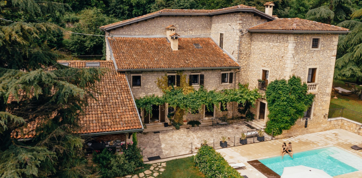 An old stone house with a red-tile roof, surrounded by trees and vines, with a swimming pool and two people sitting on the pool's edge.