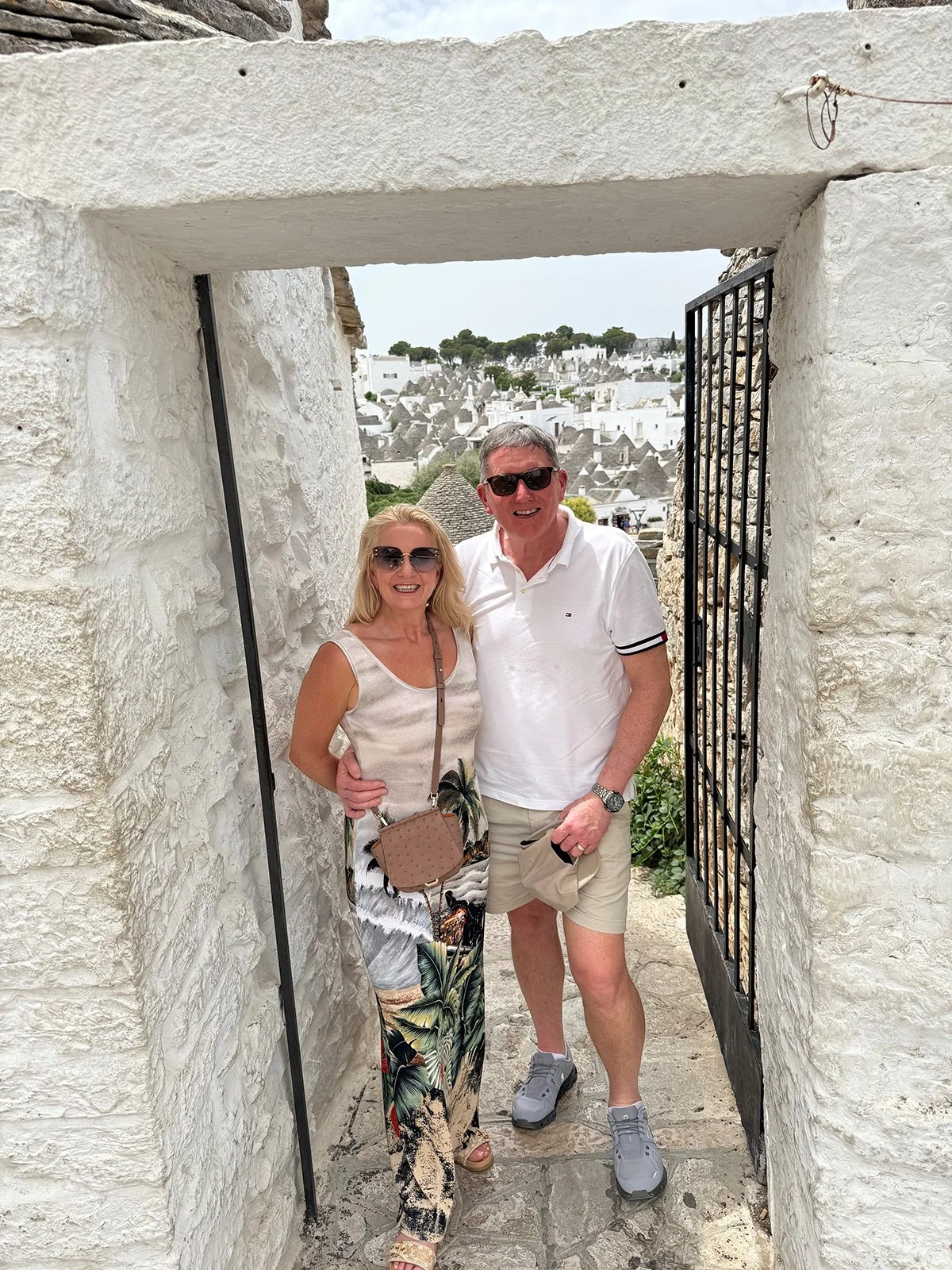 A smiling couple stands together under an arched stone doorway with a gate, with white buildings and hills in the background.
