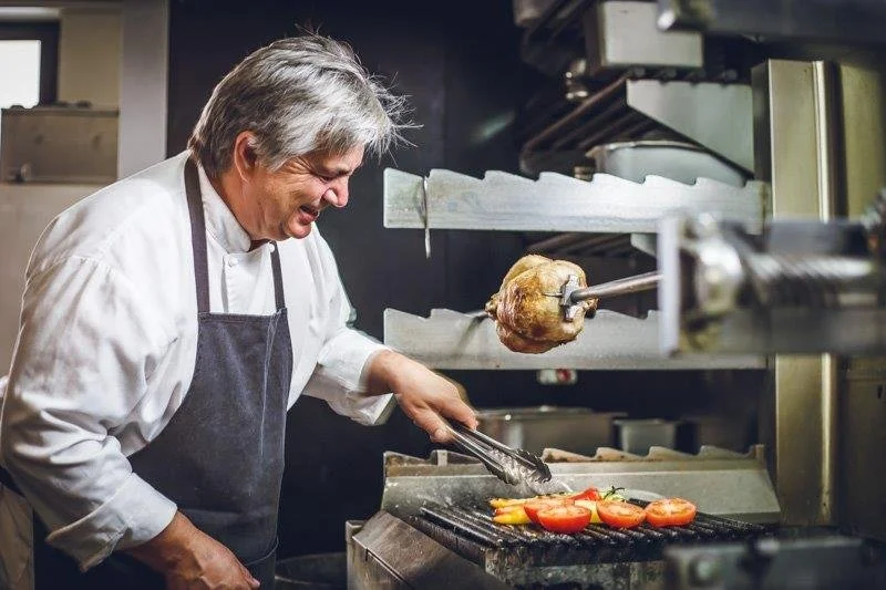 A chef grilling tomatoes and peppers while a rotating spit with meat is in the oven.