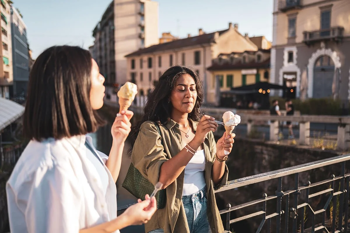 Two women standing by a bridge holding ice cream cones in an urban setting during sunset.