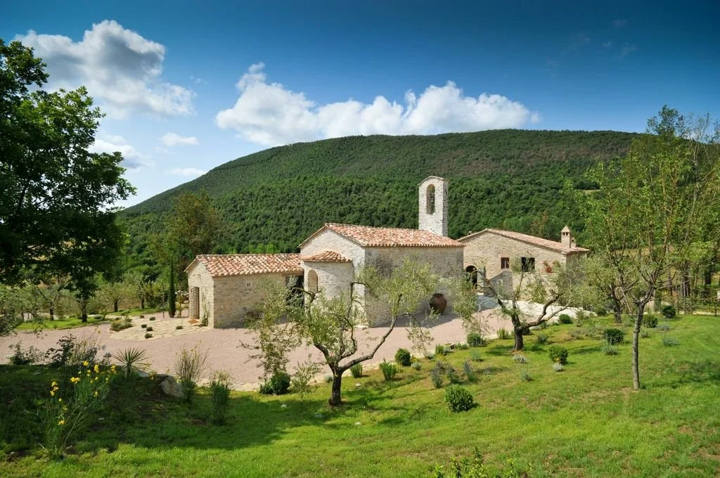 A stone church with a red-tiled roof and a small bell tower, surrounded by a garden with trees and shrubs, set against a green hill and blue sky with clouds.