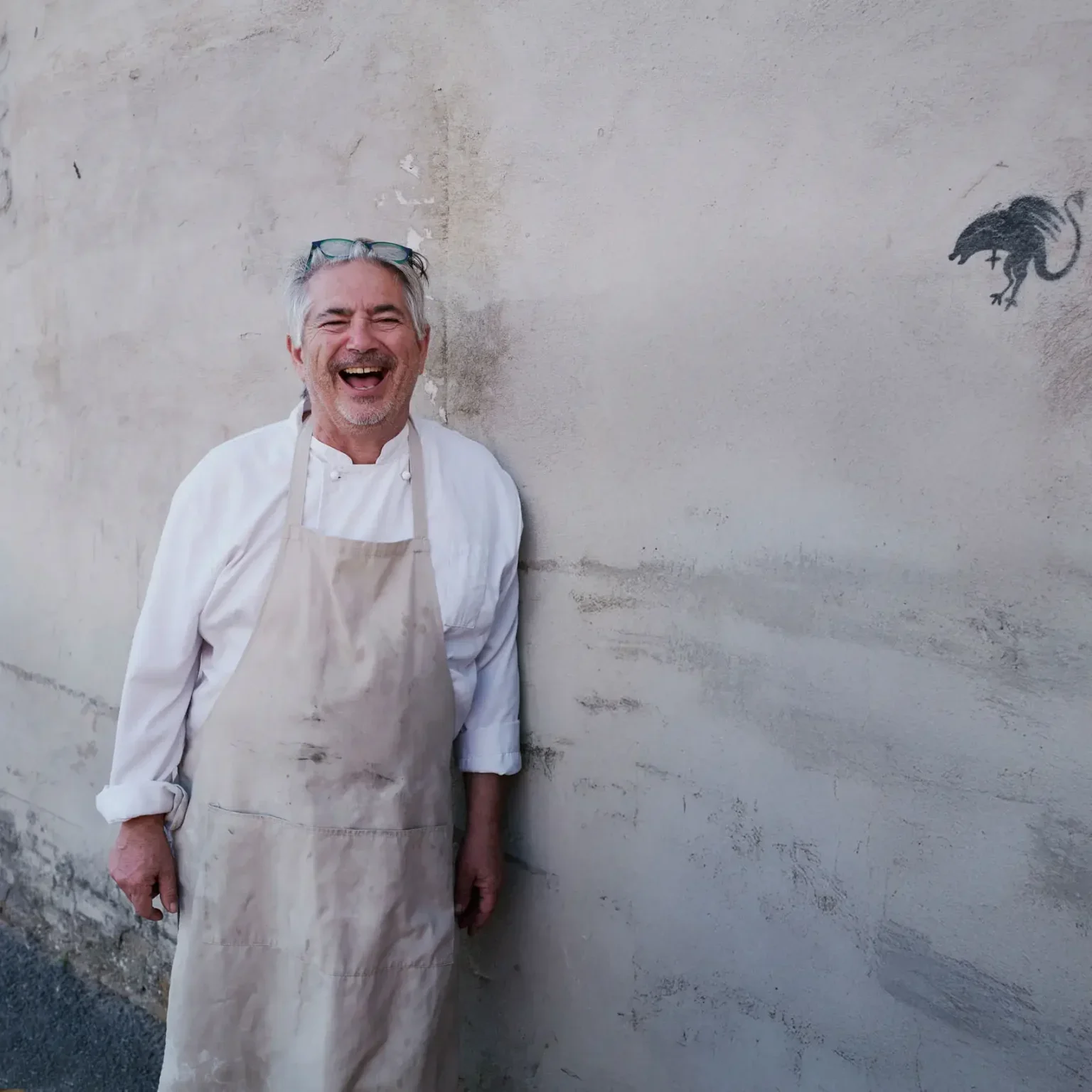 A cheerful man dressed as a chef standing against a weathered wall with a small black graffiti of a cartoonish rat or mouse in the upper right corner.