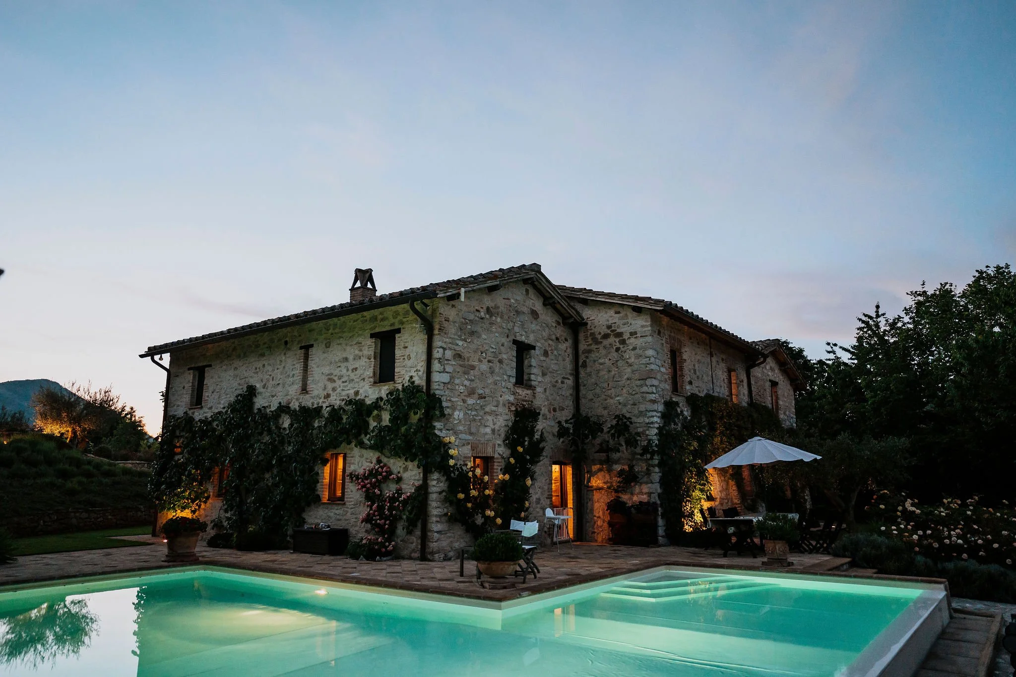 A stone house with vine-covered walls, lit windows, and outdoor furniture around a swimming pool at dusk.