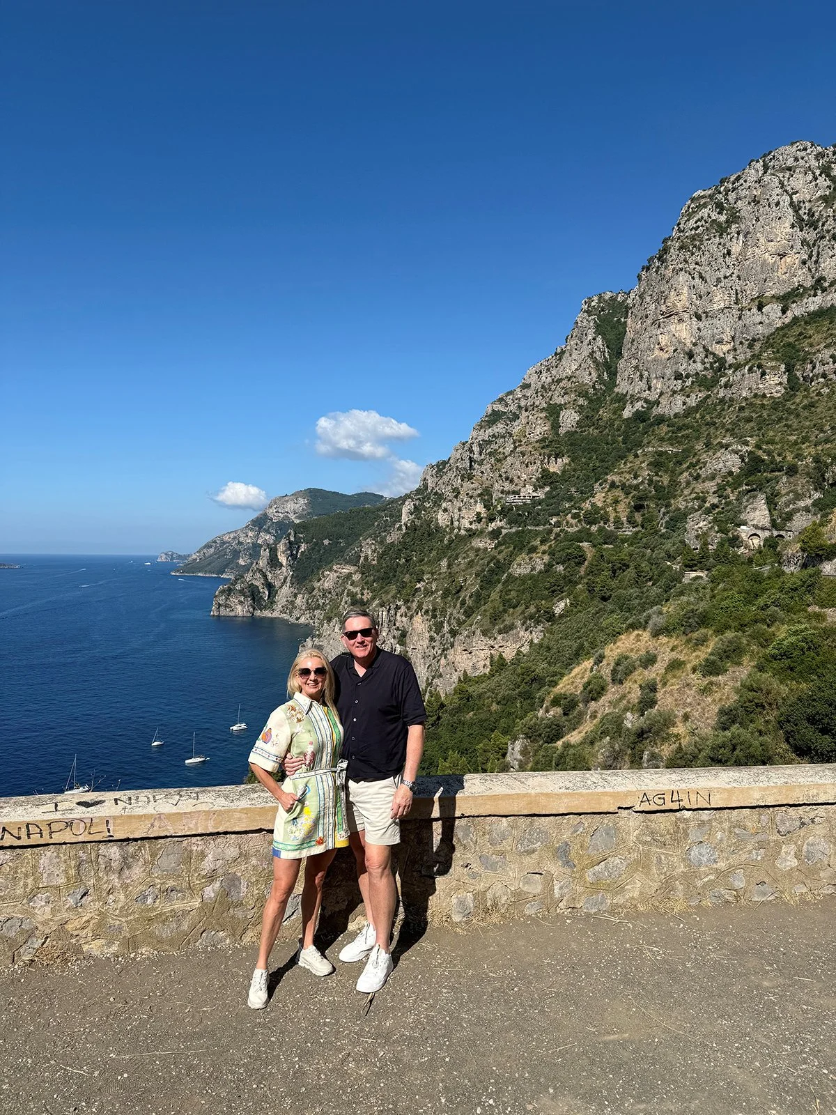 A couple standing on a stone viewpoint with a scenic coastal background, including cliffs, ocean, sailboats, and a clear blue sky.