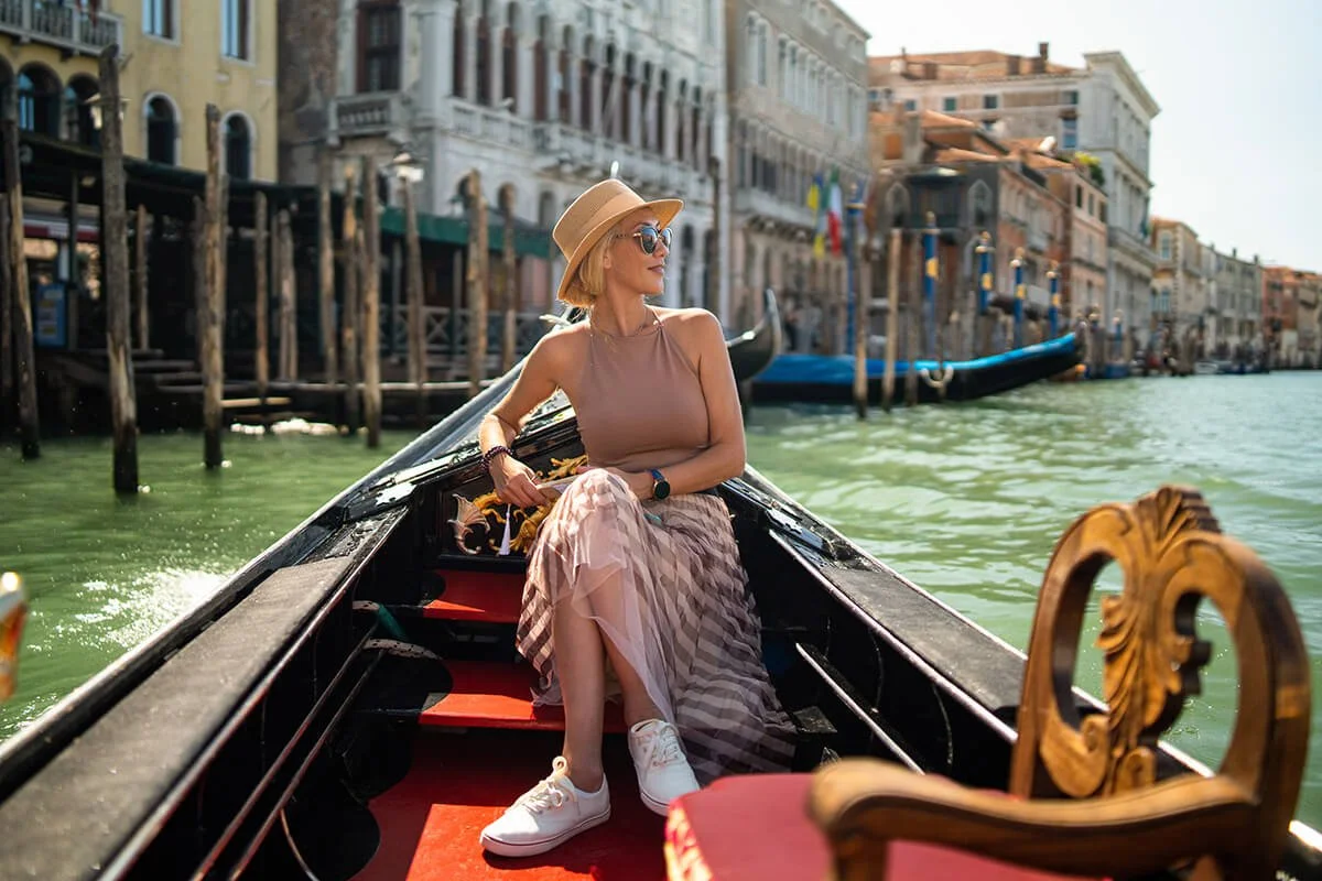 A woman in a tan sleeveless top, long striped skirt, white sneakers, wide-brimmed hat, and sunglasses sits in a gondola on a canal in Venice, Italy, with historic buildings along the water and other gondolas in the background.