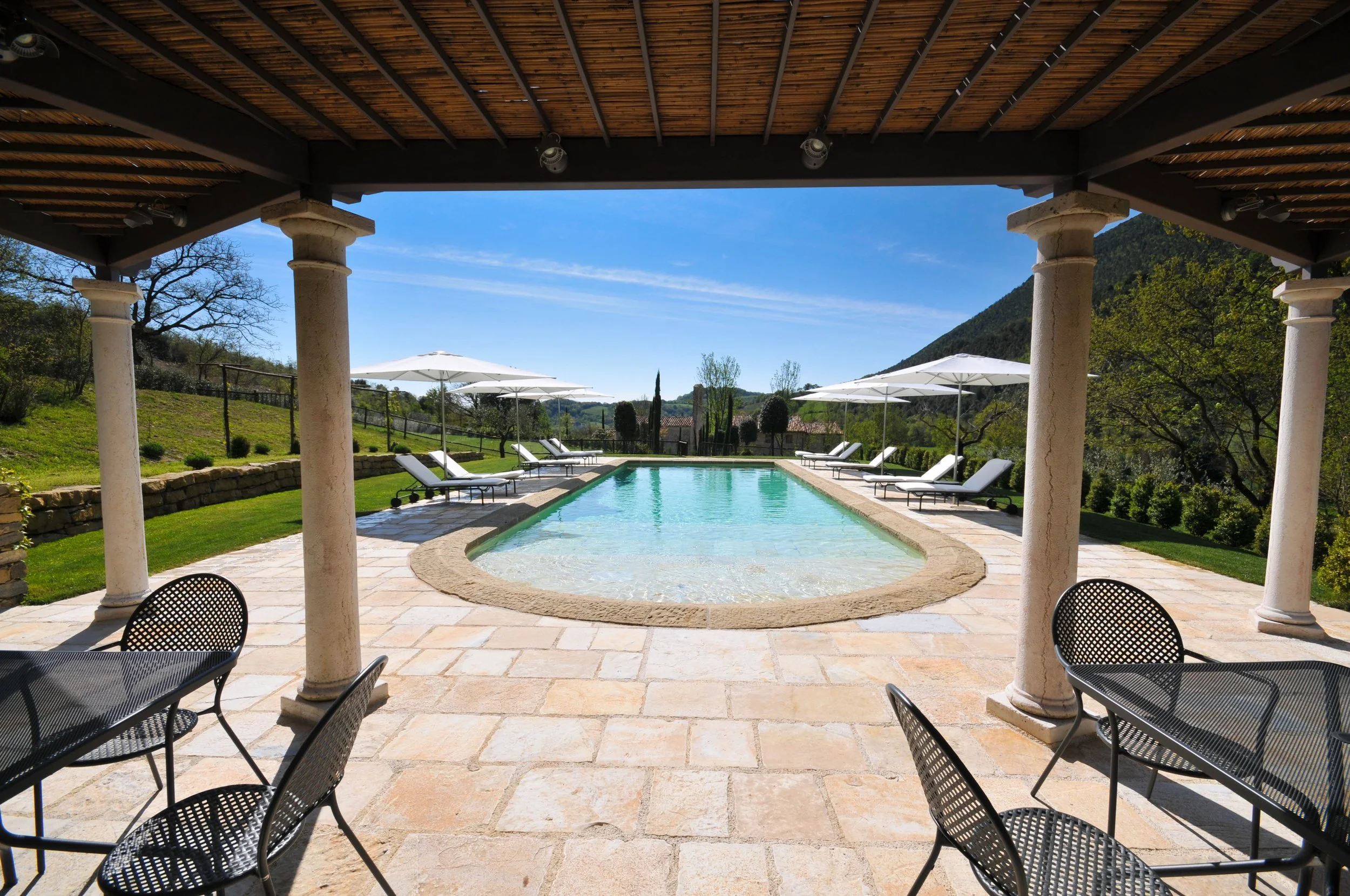 View of an outdoor swimming pool with poolside chairs and umbrellas, surrounded by green grass, trees, and distant mountains under a clear blue sky.