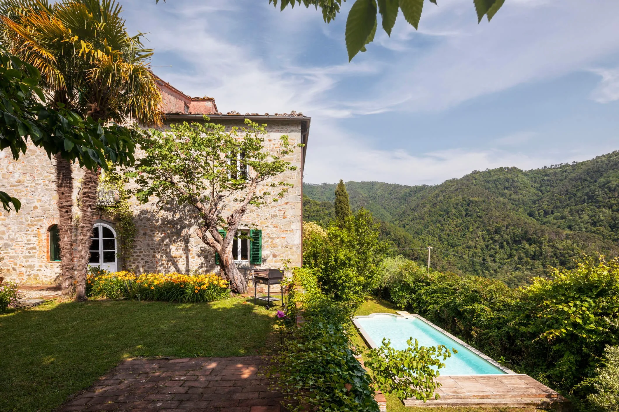 Stone house with lush trees, a swimming pool, and green hills in the background.